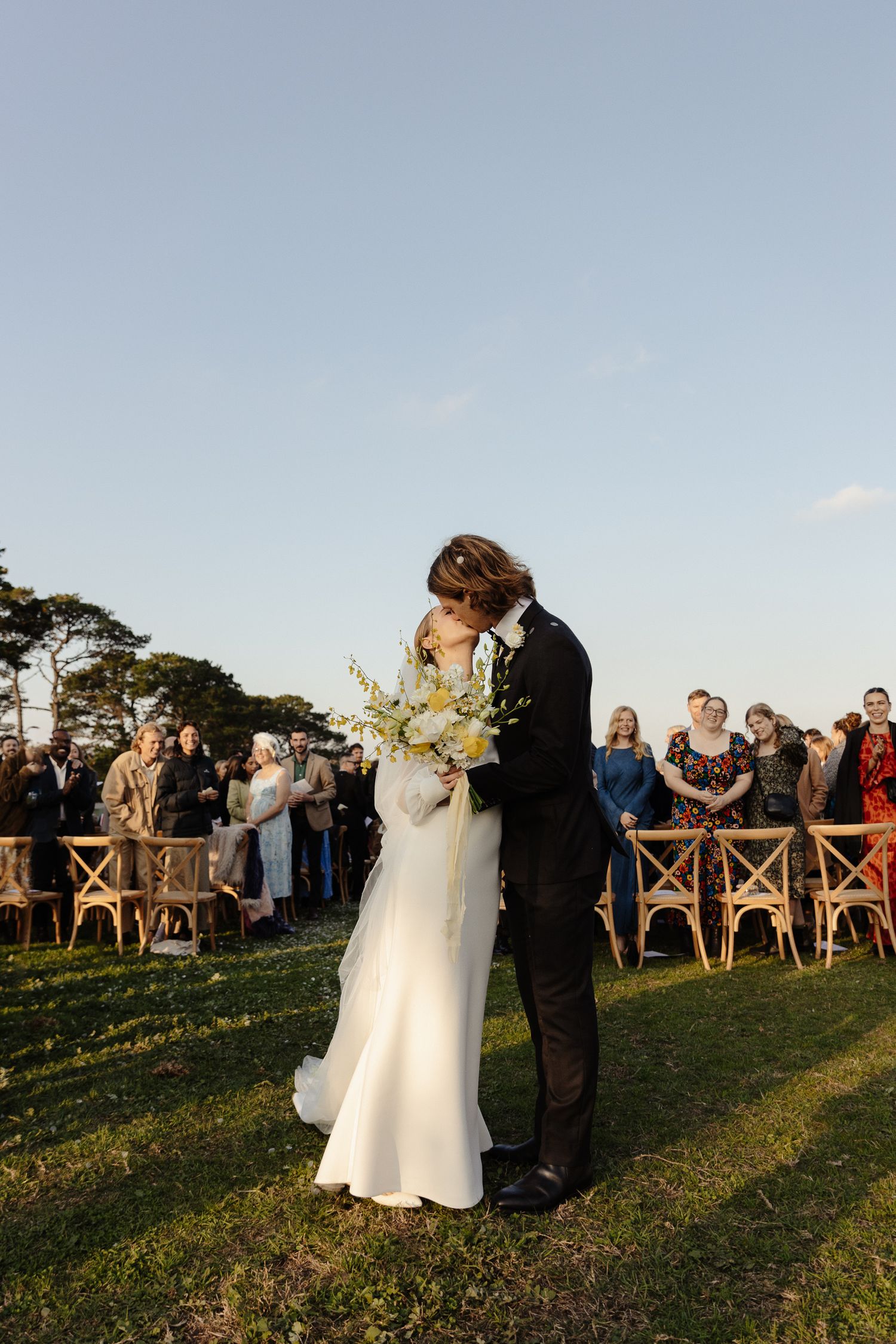 A wedding ceremony takes place outdoors with guests watching the couple share an intimate moment.
