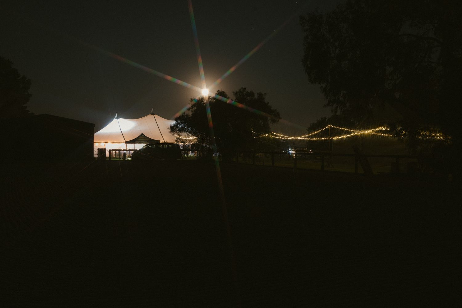White tent illuminated at night under bright spotlight with dark silhouettes of trees against starry sky.