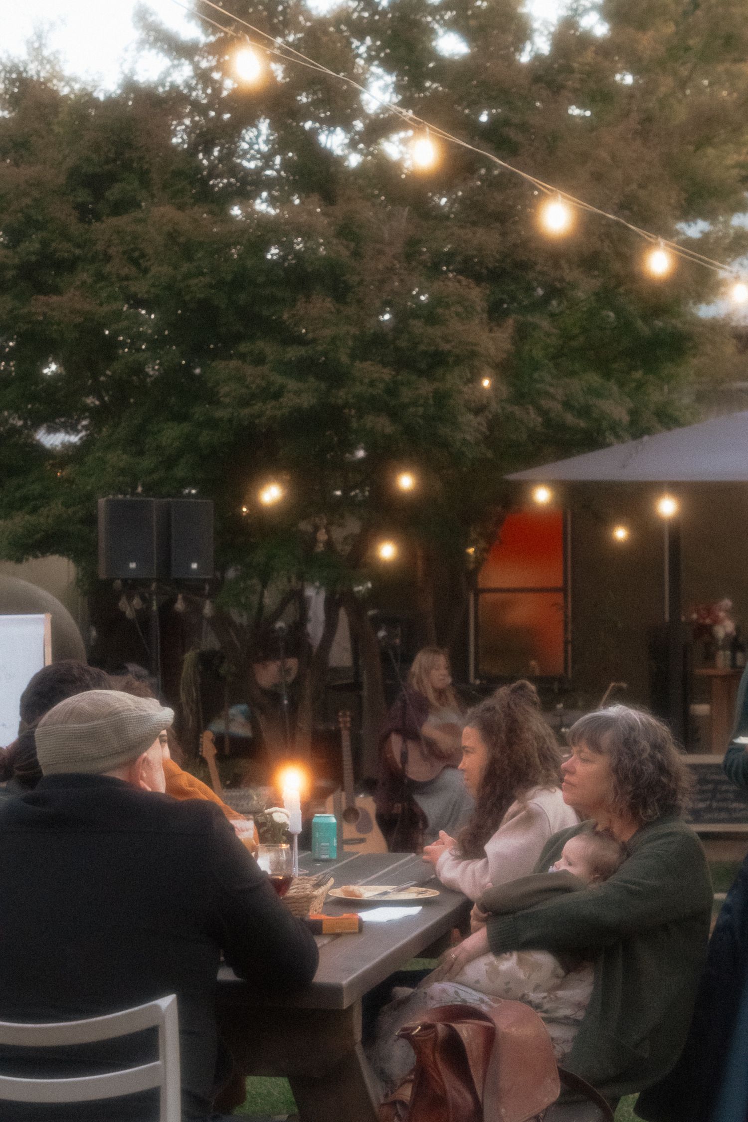 People gather at an outdoor dining table under glowing string lights strung between trees at dusk.