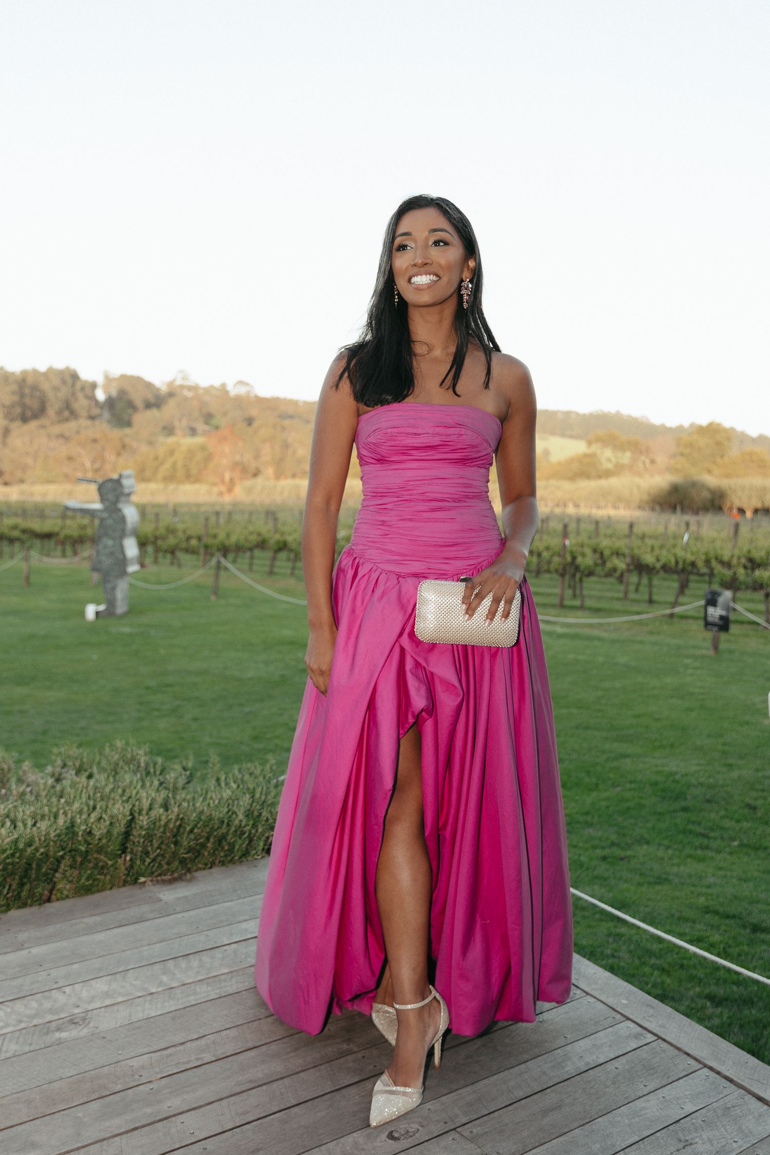 Bridesmaid in vibrant pink dress poses outdoors at vineyard wedding venue on wooden deck.