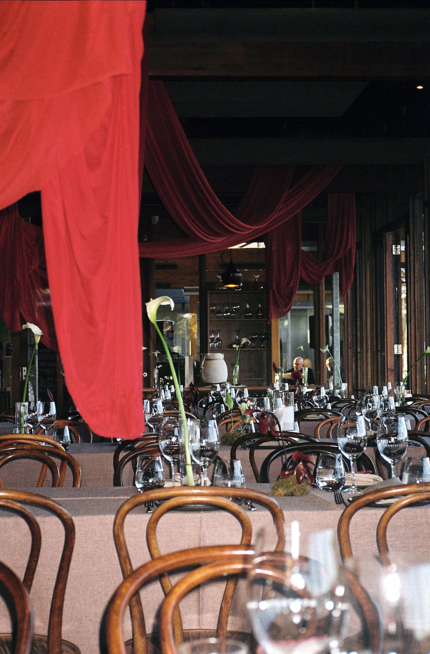 Red curtains drape elegantly in an empty restaurant dining room with wooden chairs and white tablecloths.