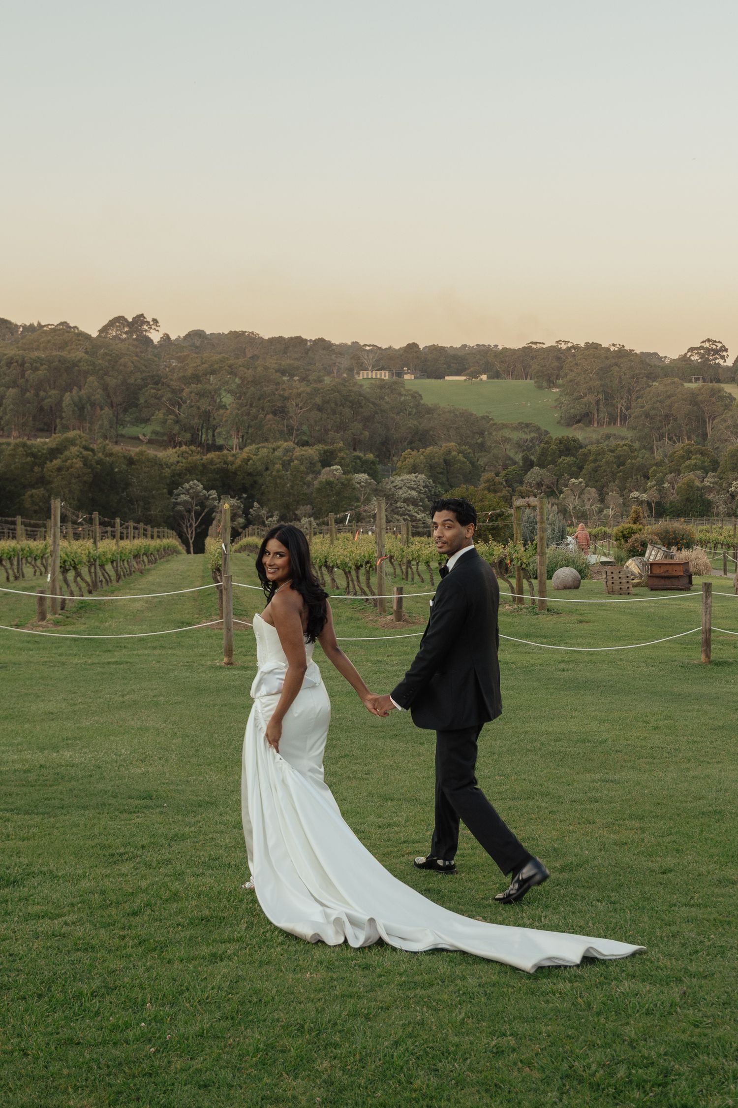 Playful couple poses in formal attire on a grassy field at a vineyard during sunset.