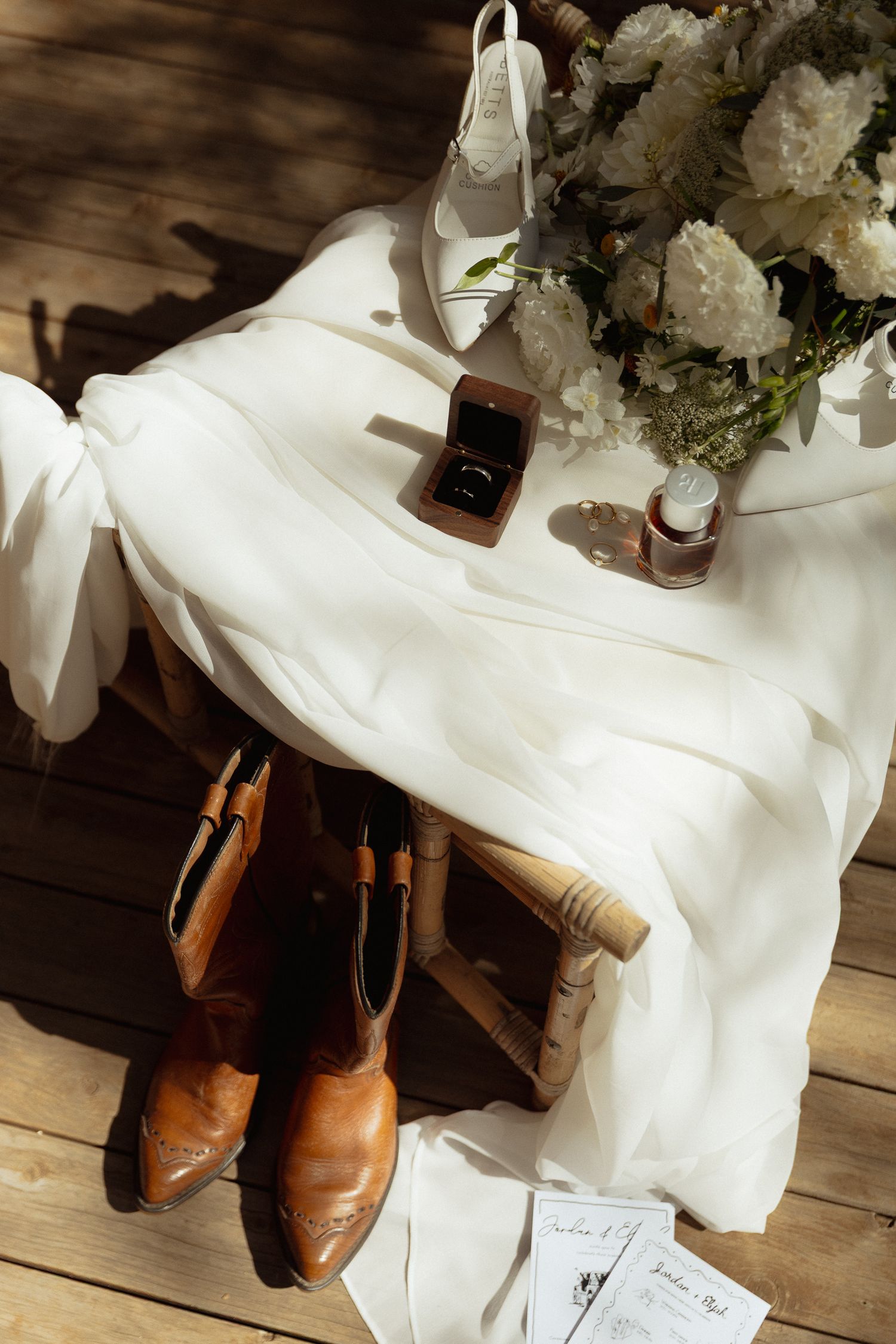 White wedding dress, brown leather boots, and white floral bouquet arranged on wooden floor with sunlight.