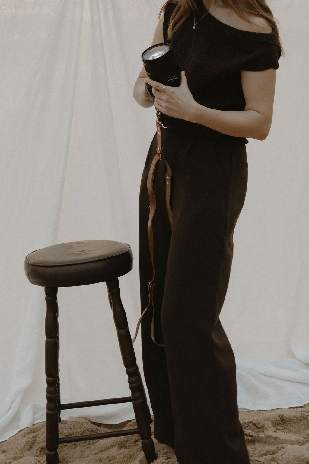 A person in a black dress stands next to a dark wooden chair against a white backdrop in a studio setting.