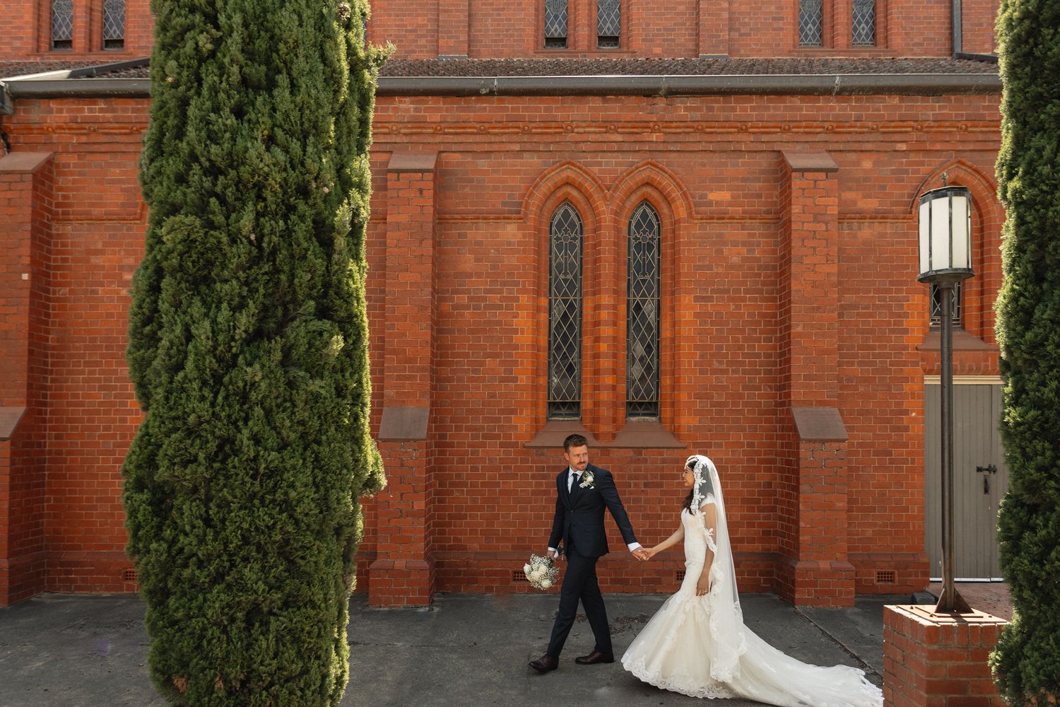 A couple walks together outside a red brick church with towering cypress trees on their wedding day.