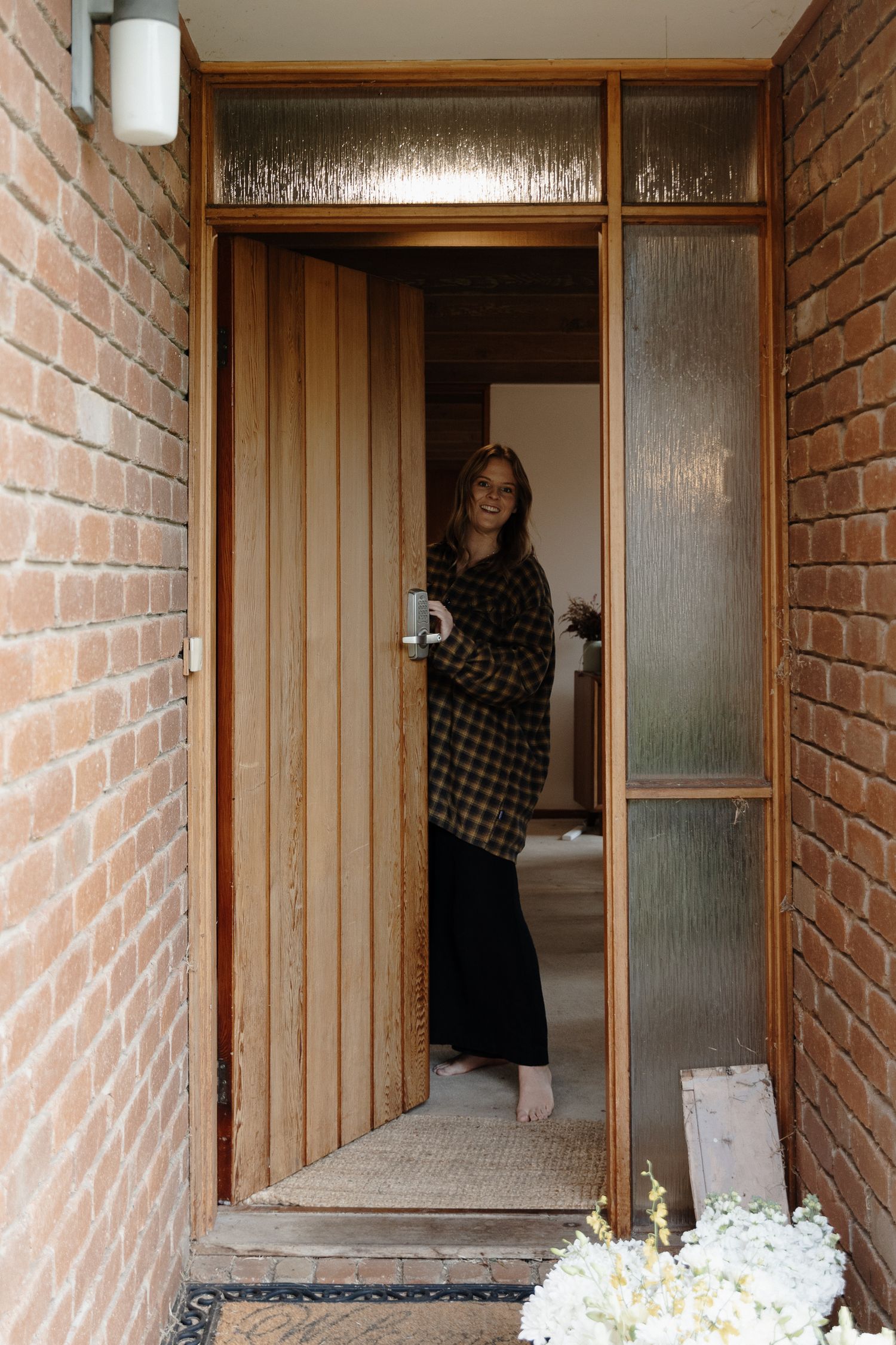 Person in plaid coat stands in a brick entryway with wooden door and textured glass panel.