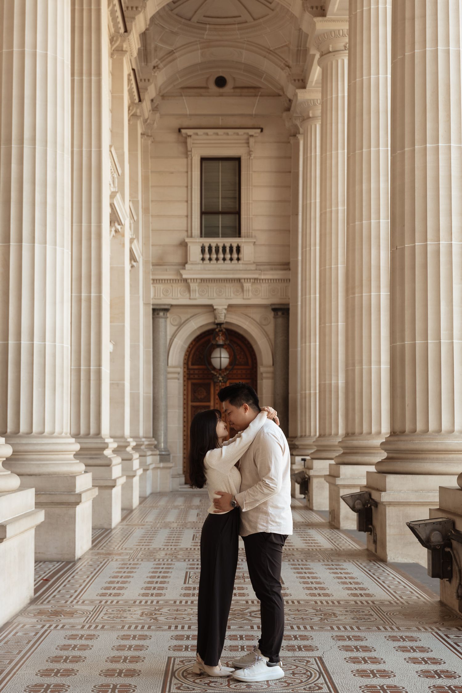 Couple embraces in a grand marble hallway with classical columns and ornate architectural details.