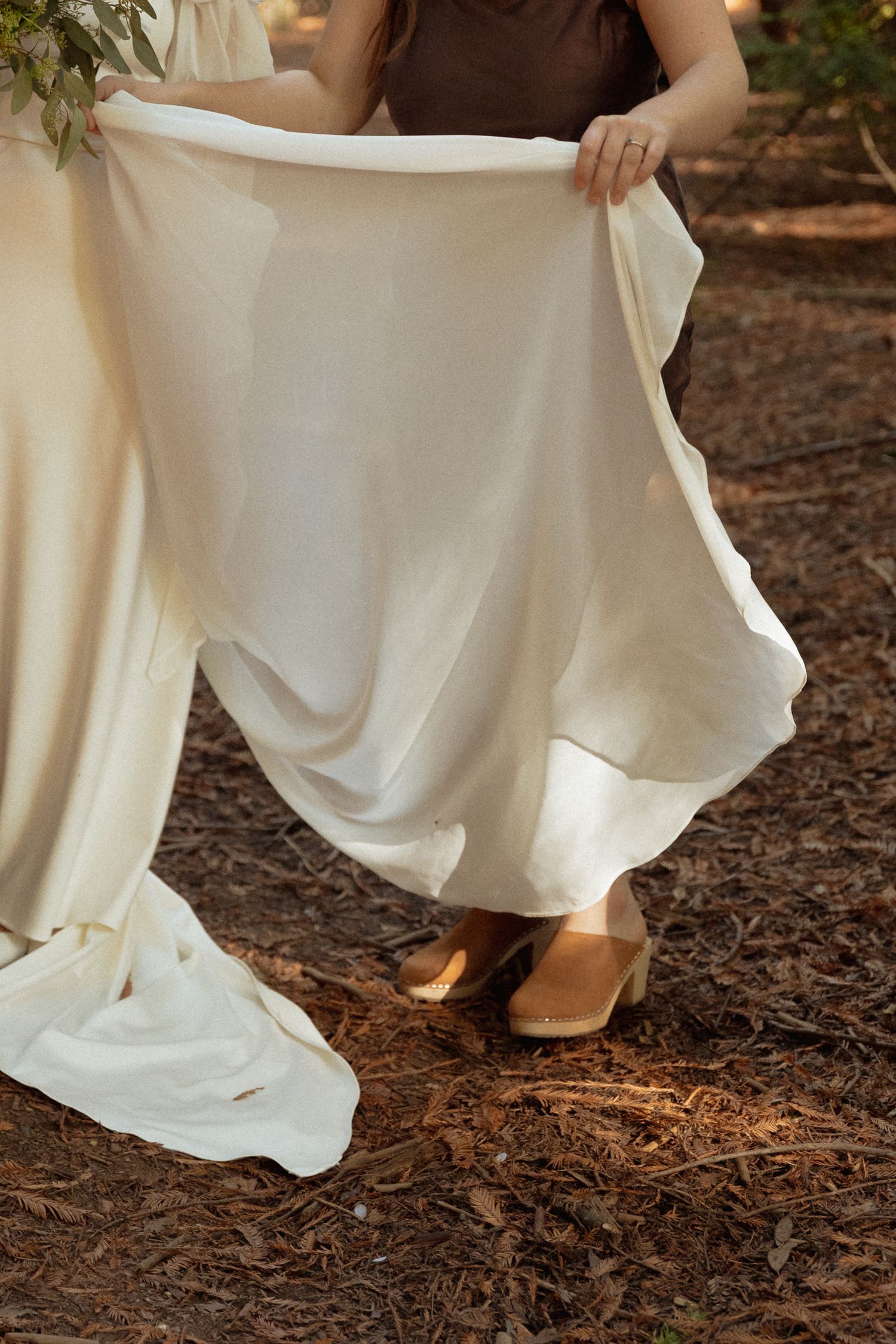 Close-up view of flowing white dress hem and brown leather shoes on forest ground covered in pine needles.
