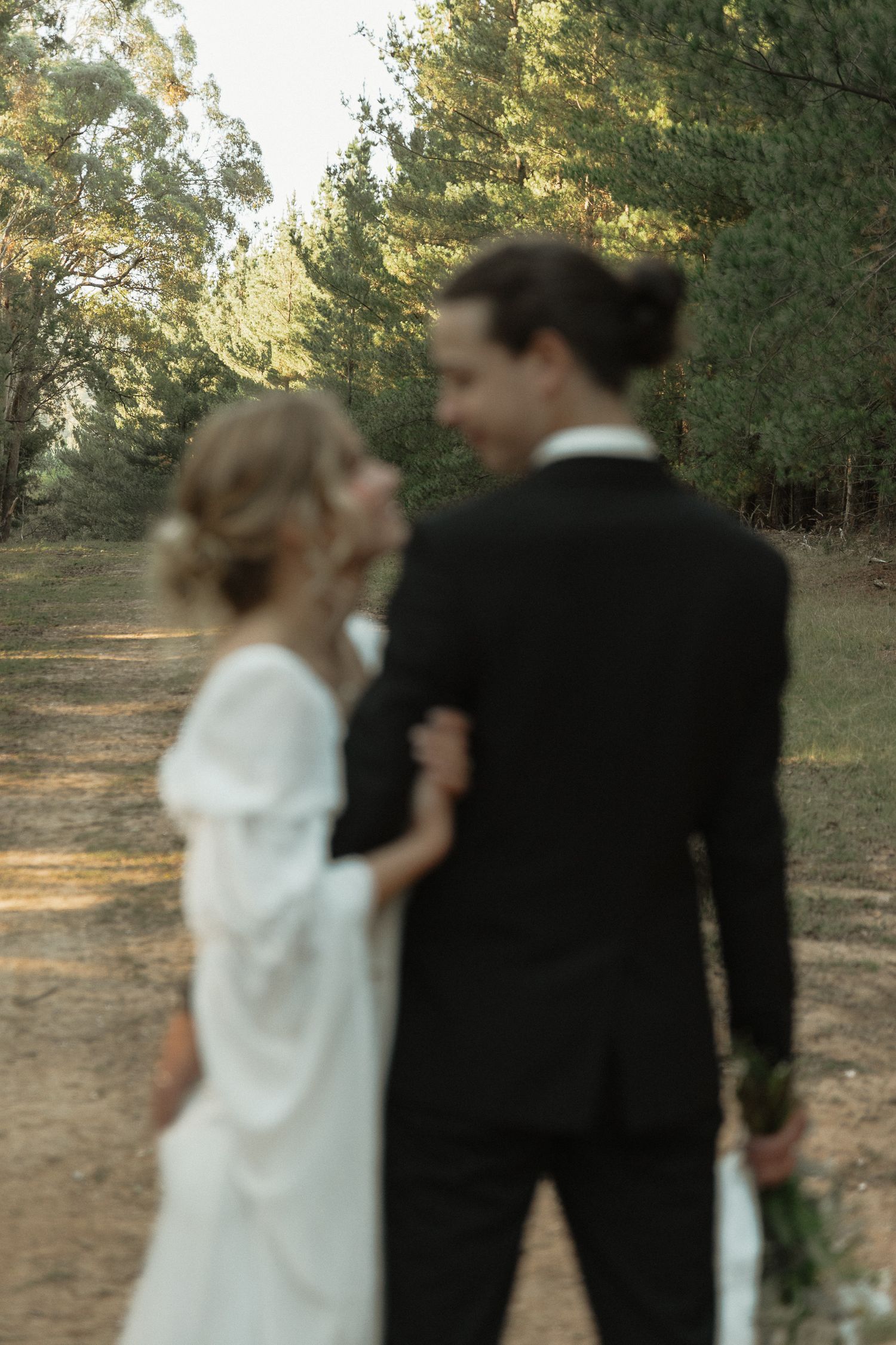 A romantic wedding couple shares an intimate moment on a sunlit forest path in formal attire.