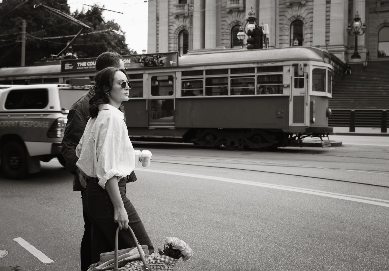 A black and white street scene shows a person waiting to cross while a vintage tram and car pass by in front of a building.