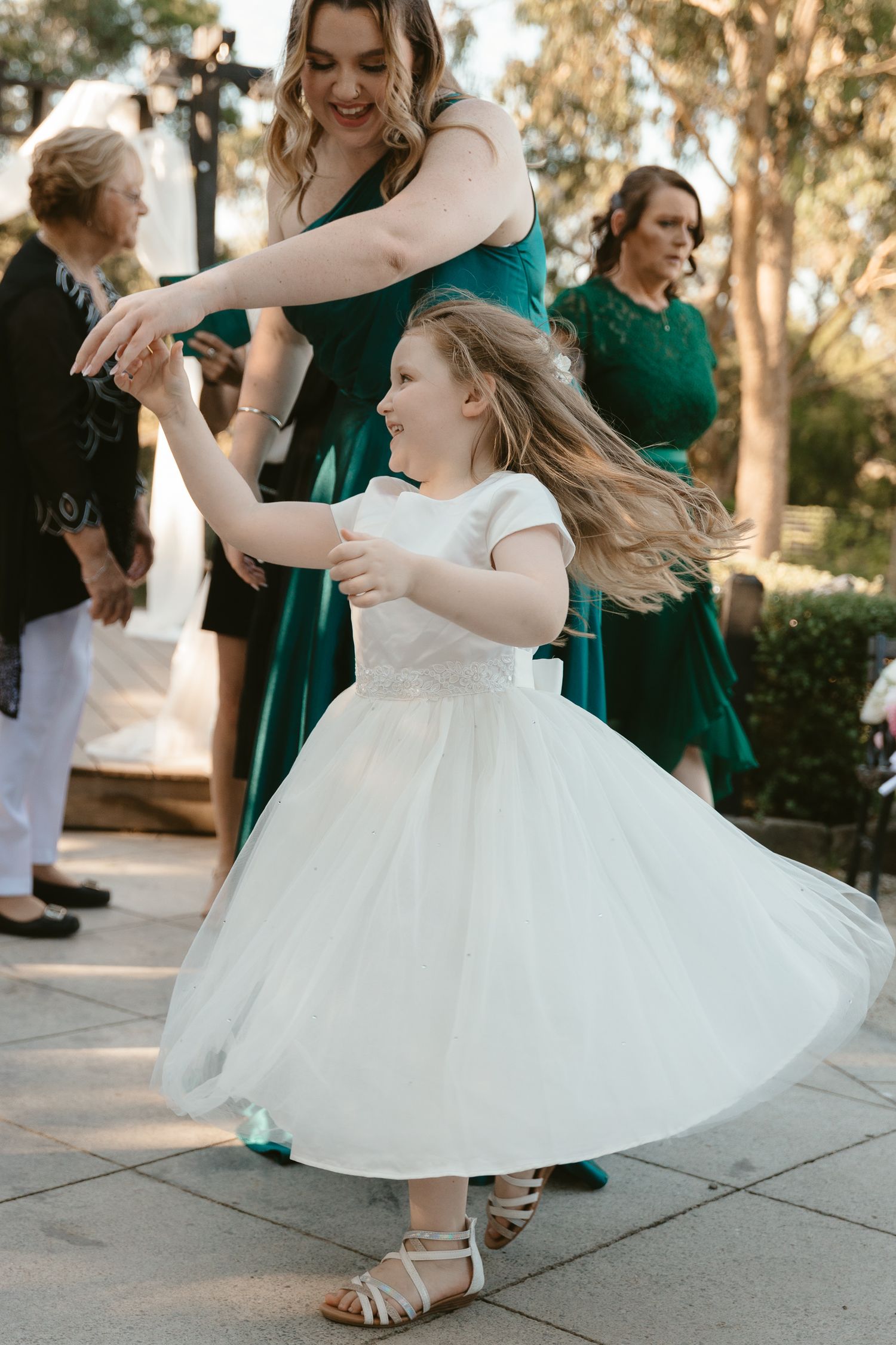 Flower girl in white dress twirling and dancing with bridesmaid in emerald green.