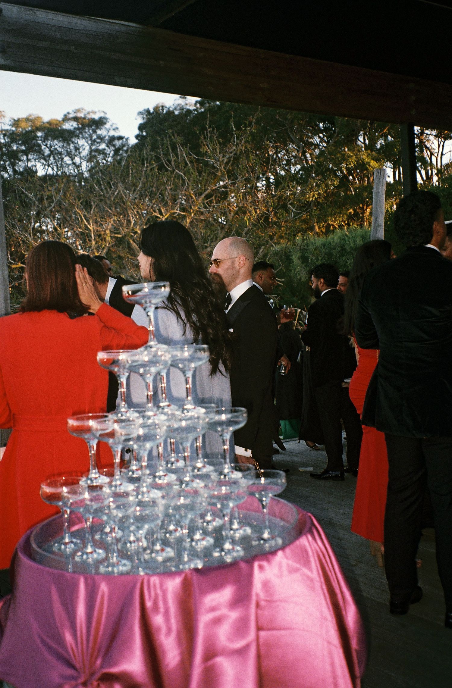Wedding reception party with champagne glasses arranged in pyramid formation on pink tablecloth at dusk.