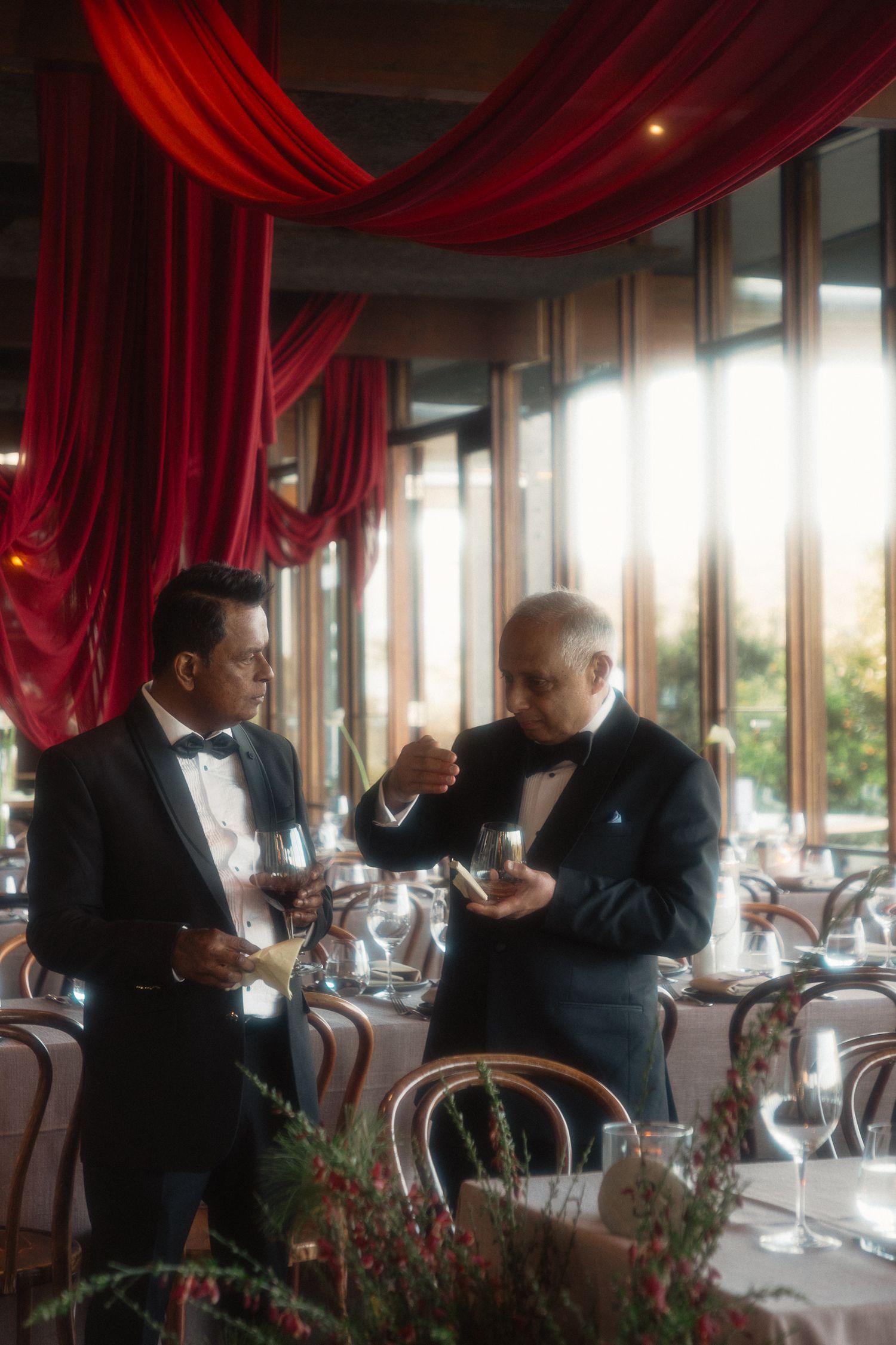 Two people in formal attire having a conversation at an elegant restaurant with red curtains and window views.