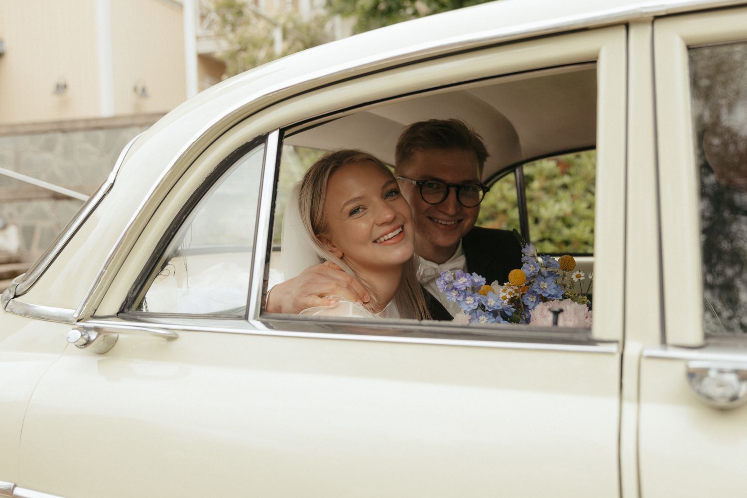 A happy couple sits in a vintage white car during their wedding celebration.