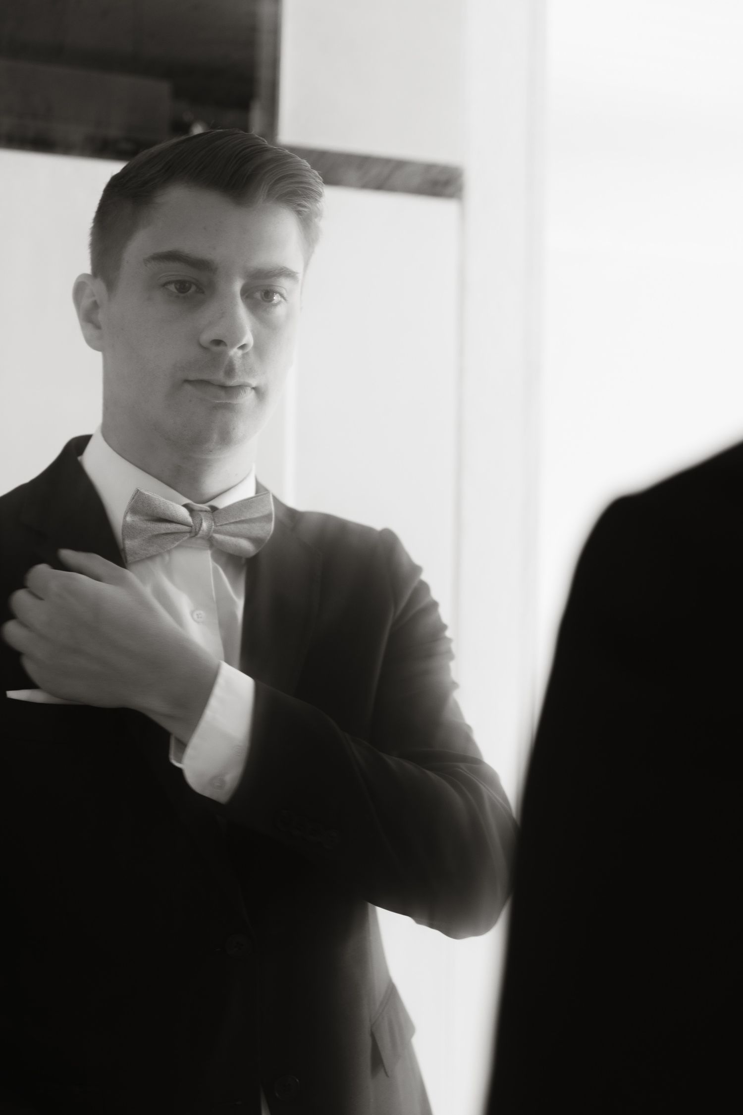 Black and white photograph of a person in formal attire with bowtie adjusting their tuxedo jacket in elegant pose.