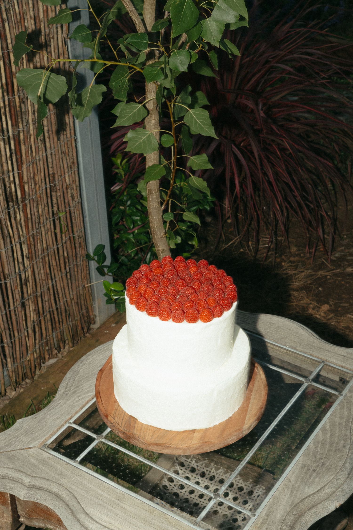 A two-tiered white cake topped with red roses sits on a wooden board against a bamboo screen backdrop with greenery.
