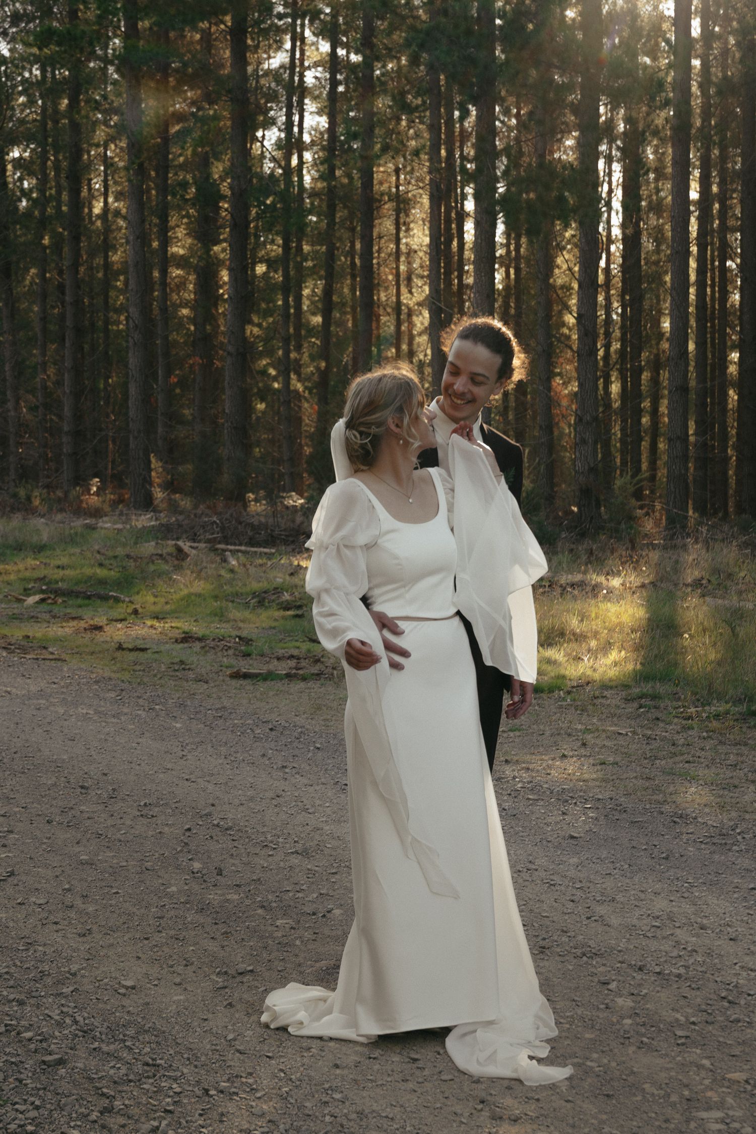 A romantic wedding moment captured on a forest path with sunlight filtering through tall pine trees.