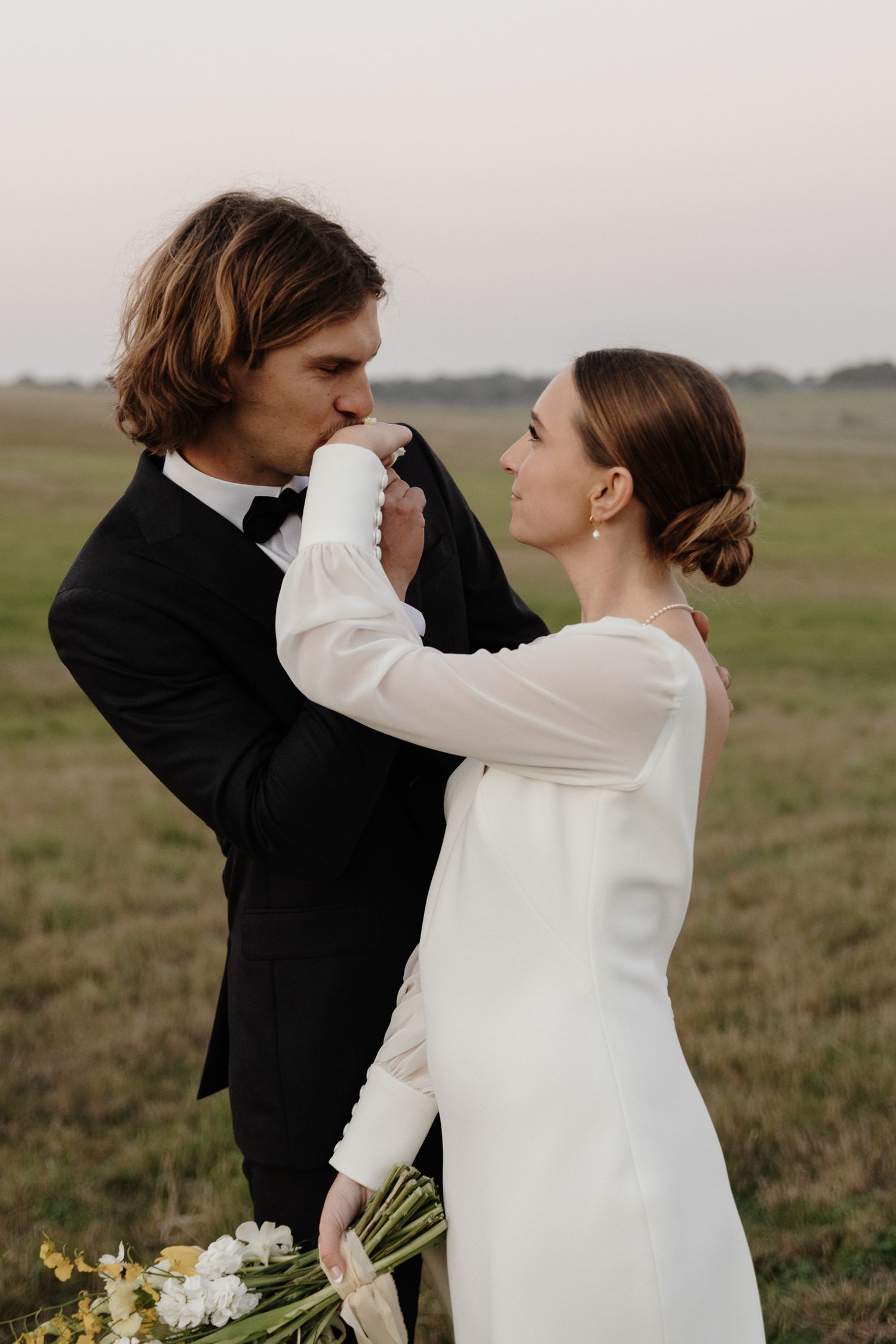 Intimate wedding portrait of couple embracing in field with bridal bouquet.
