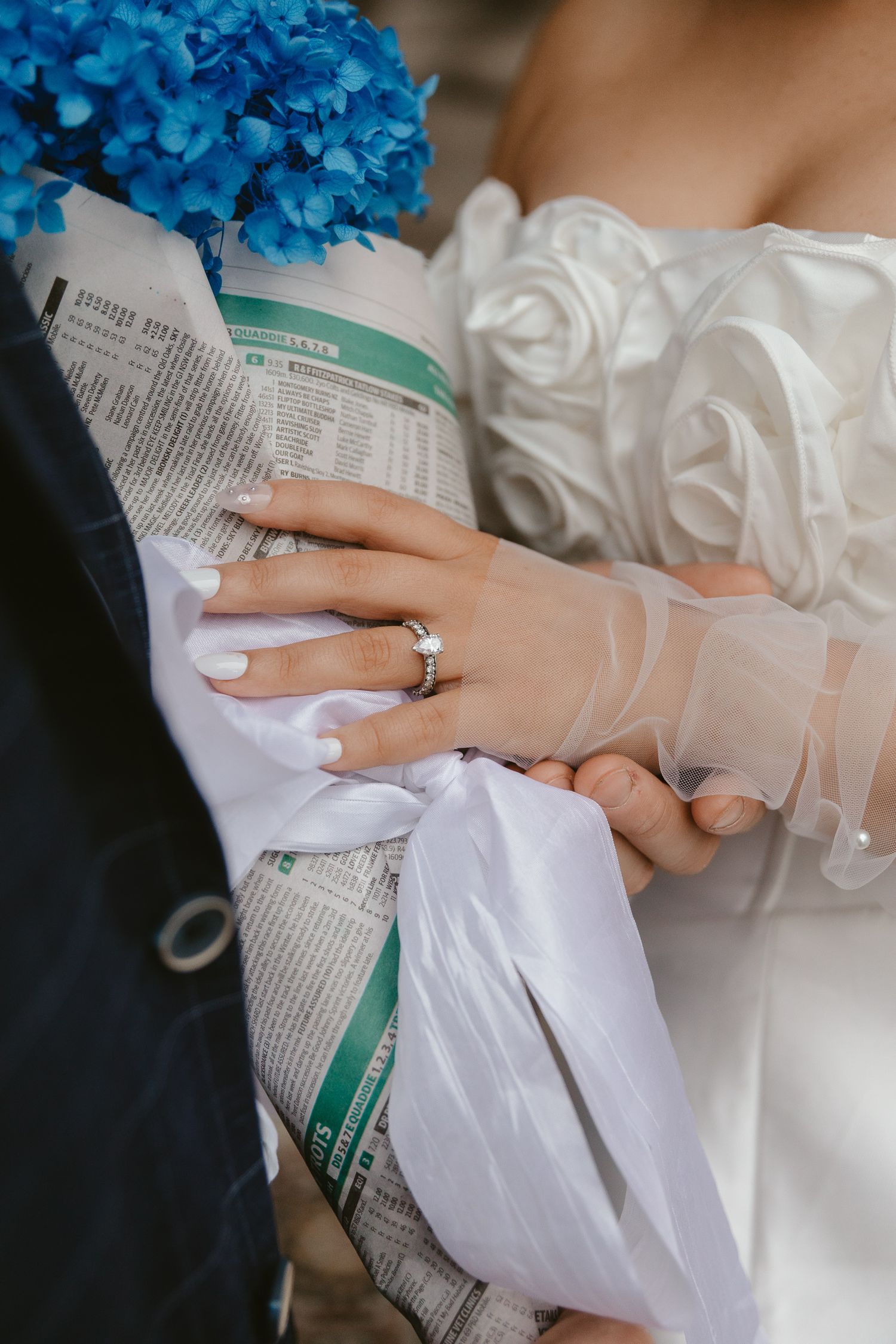 Wedding bouquet wrapped in newspaper with blue hydrangeas and bride's hands showing ring.