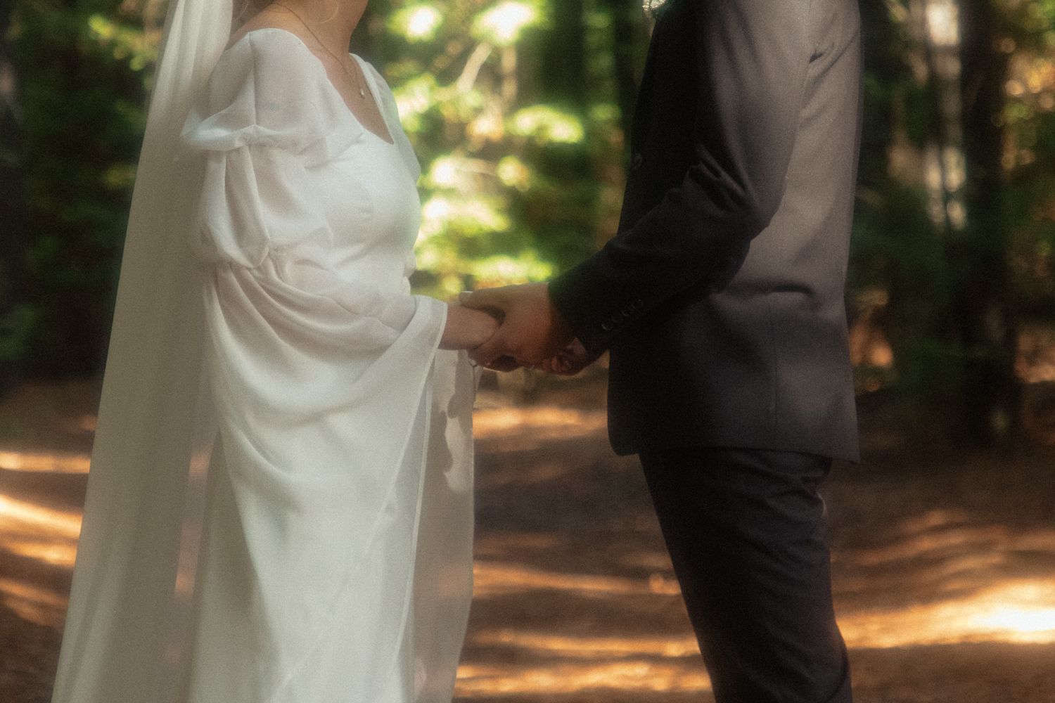 A couple holds hands in sunlit forest wearing formal wedding attire with soft focus dreamy effect.