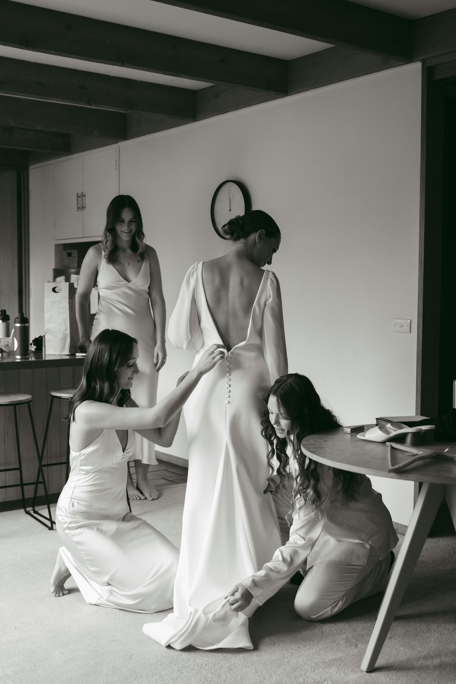 Black and white photo showing bridesmaids helping to button up a wedding dress in a minimalist room interior.