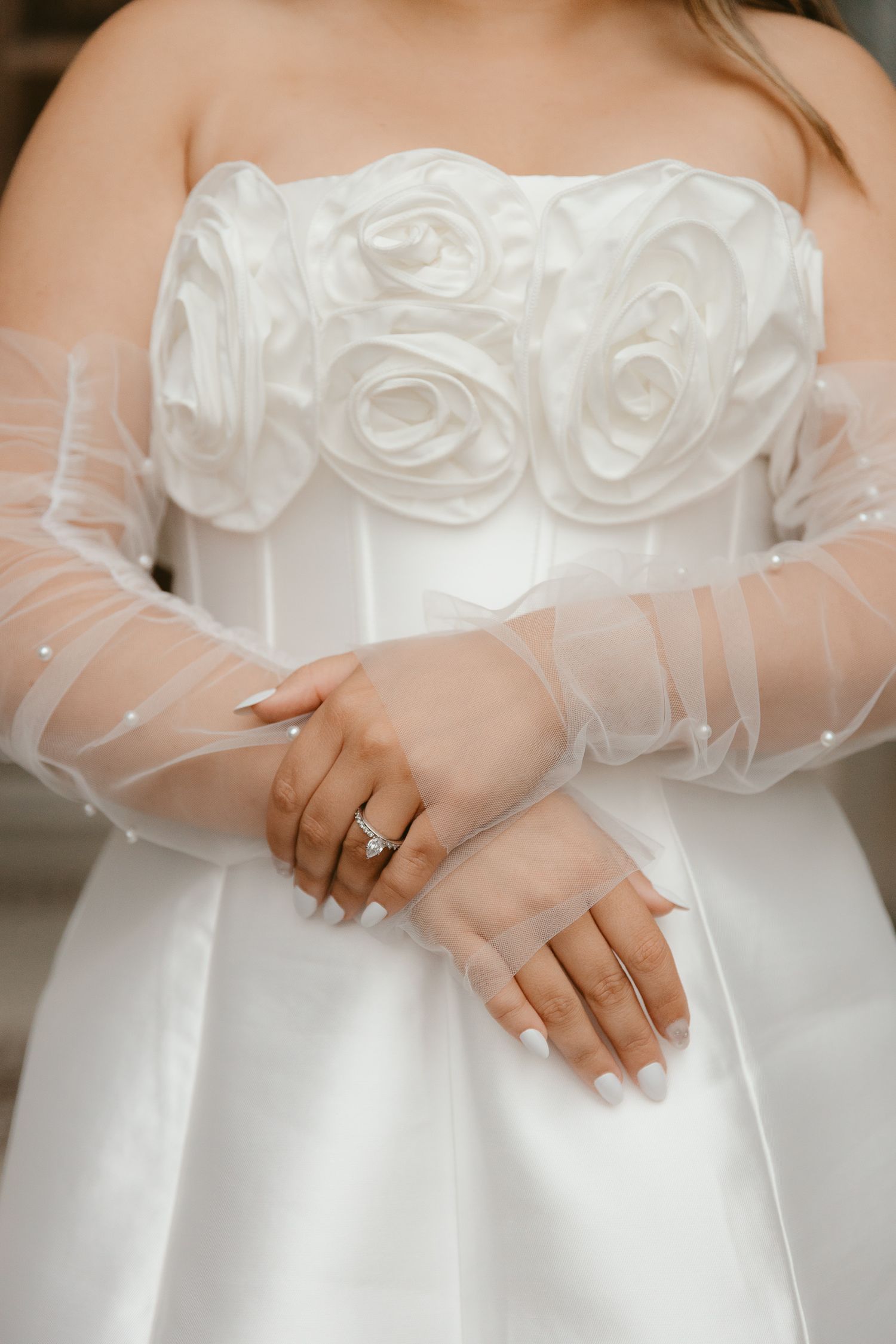Close-up of white wedding dress bodice with rose details and sheer sleeves showing wedding ring.