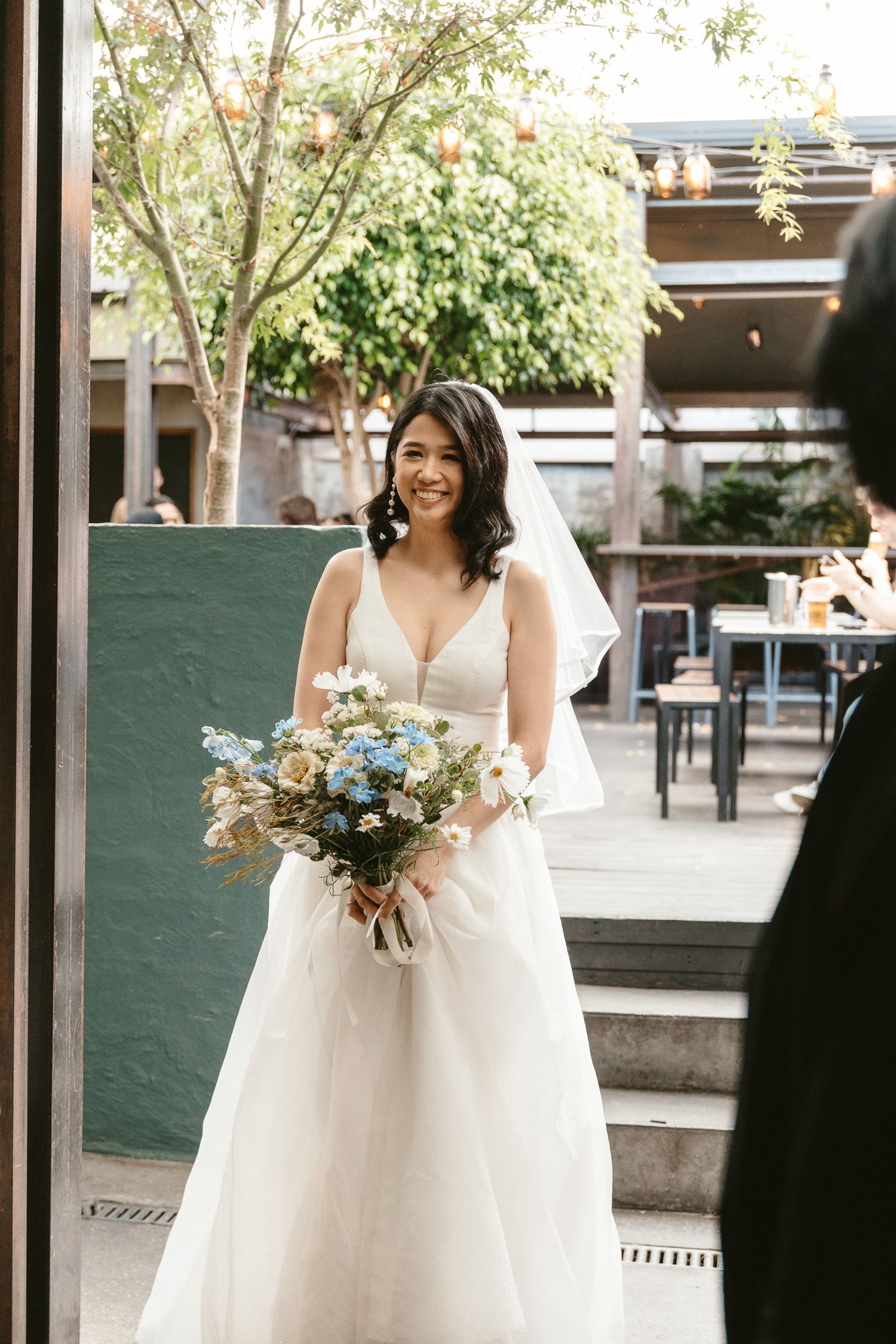 Bride in white wedding gown holding blue and white bouquet on outdoor patio.
