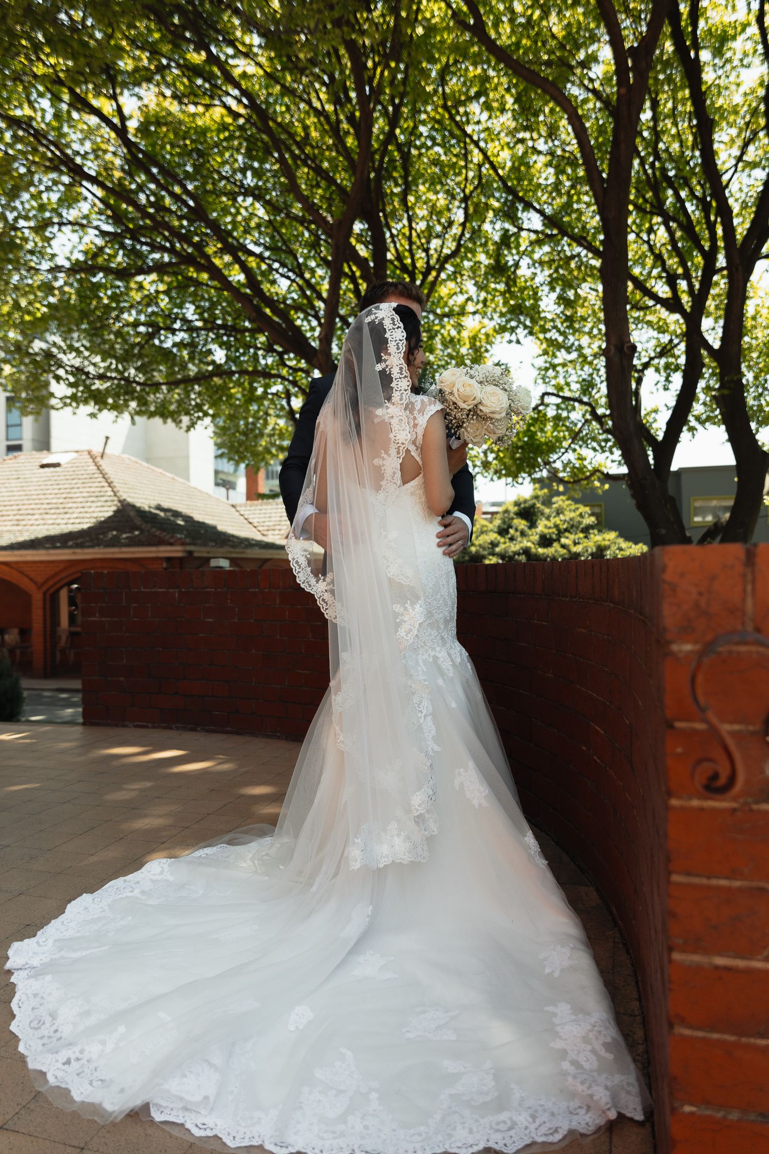 A bride in a flowing lace gown with long train poses against brick and trees.