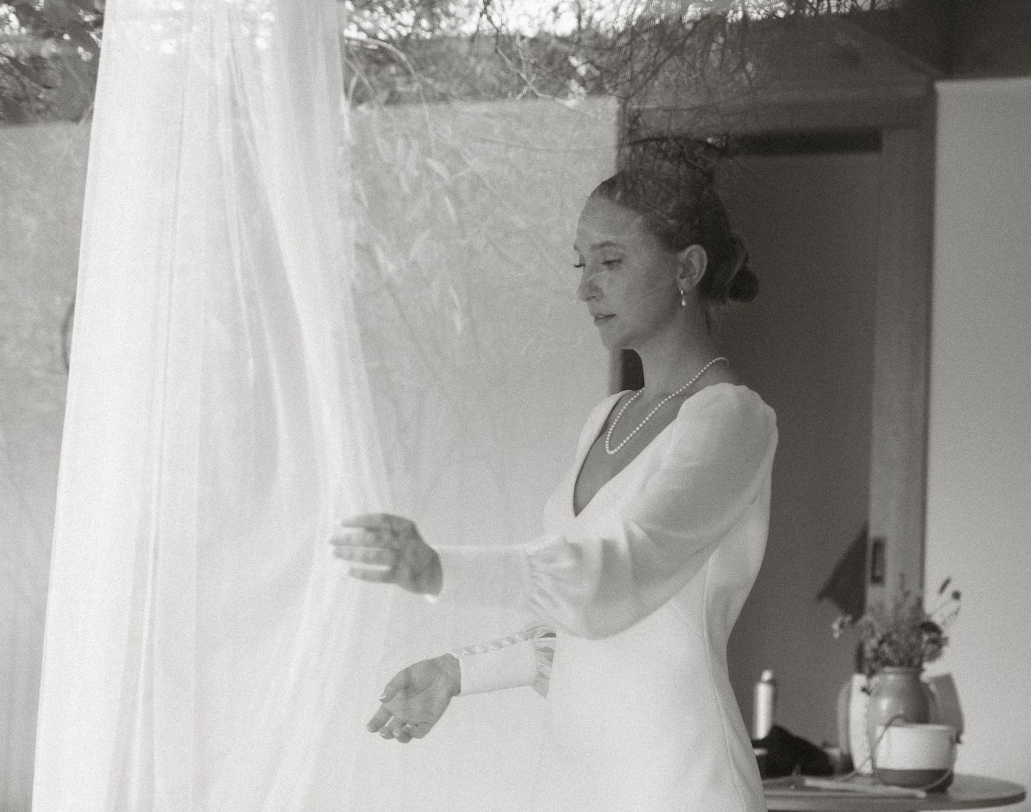 Black and white image of bride adjusting wedding veil in sunlit room.