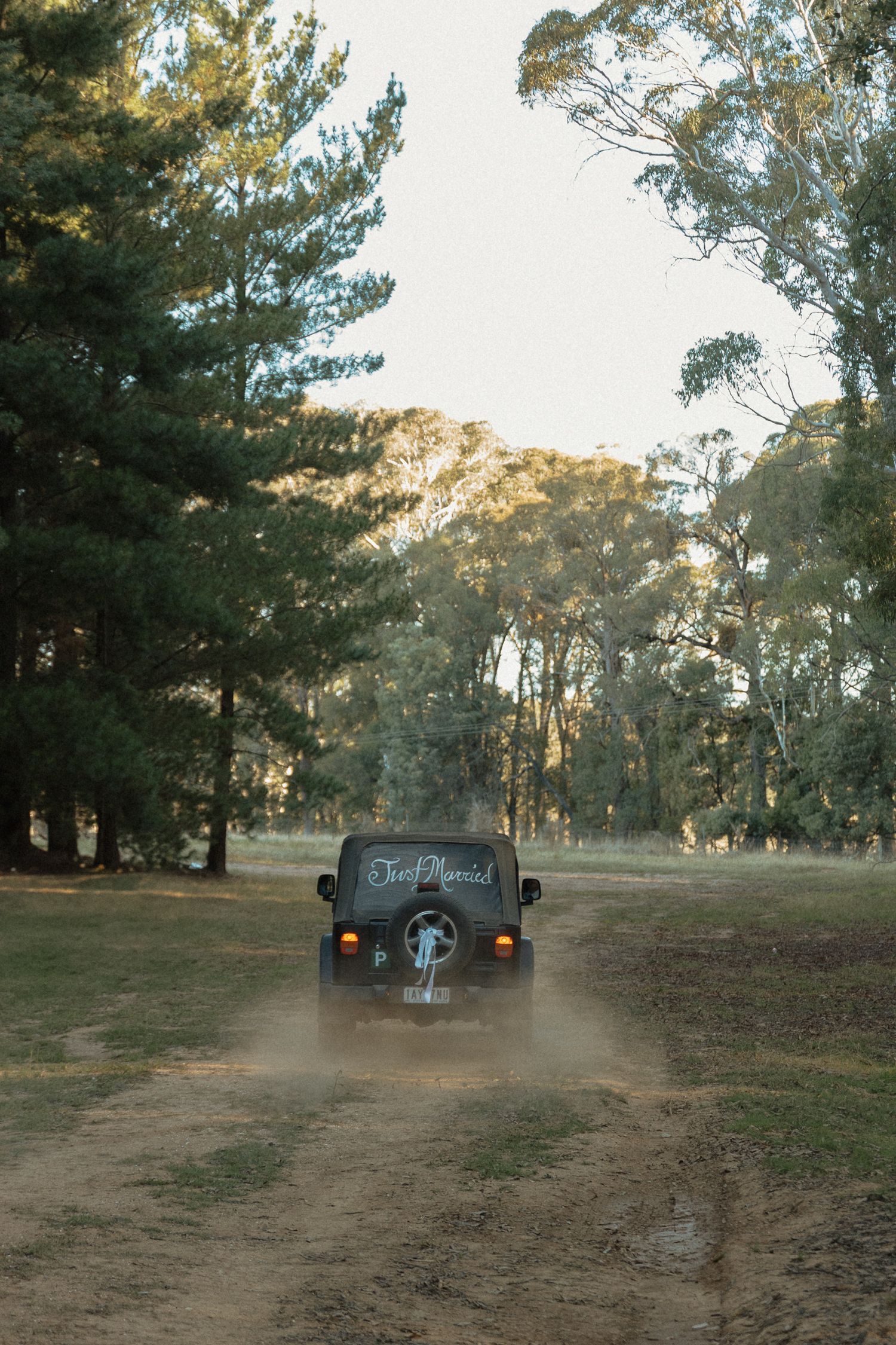 A vintage car kicks up dust while driving down a dirt road through a scenic forest at sunset.
