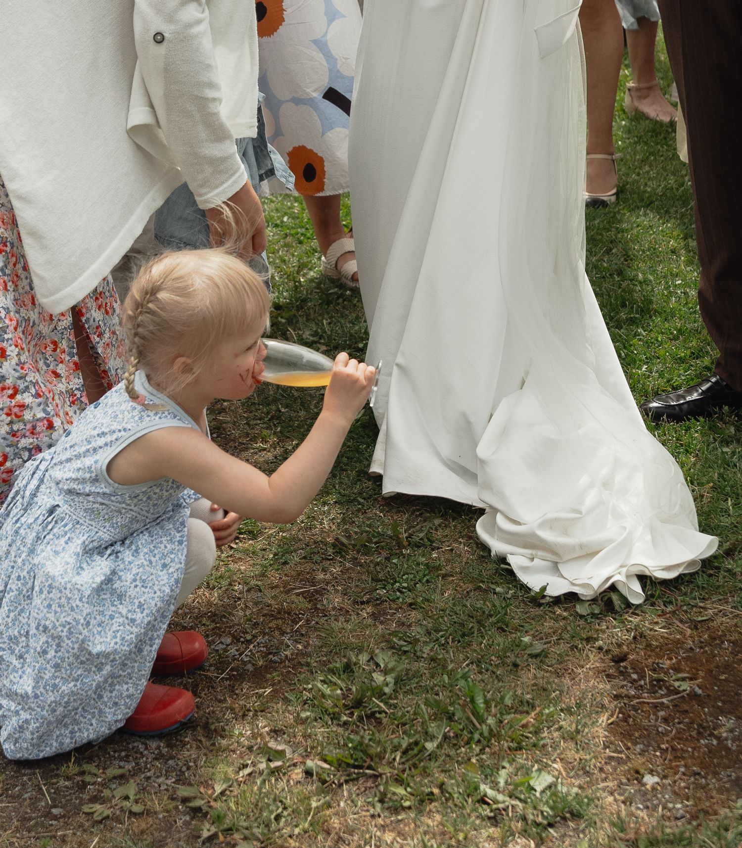 A small child in a blue dress crouches down to draw on a white wedding dress hem with a marker on grass.