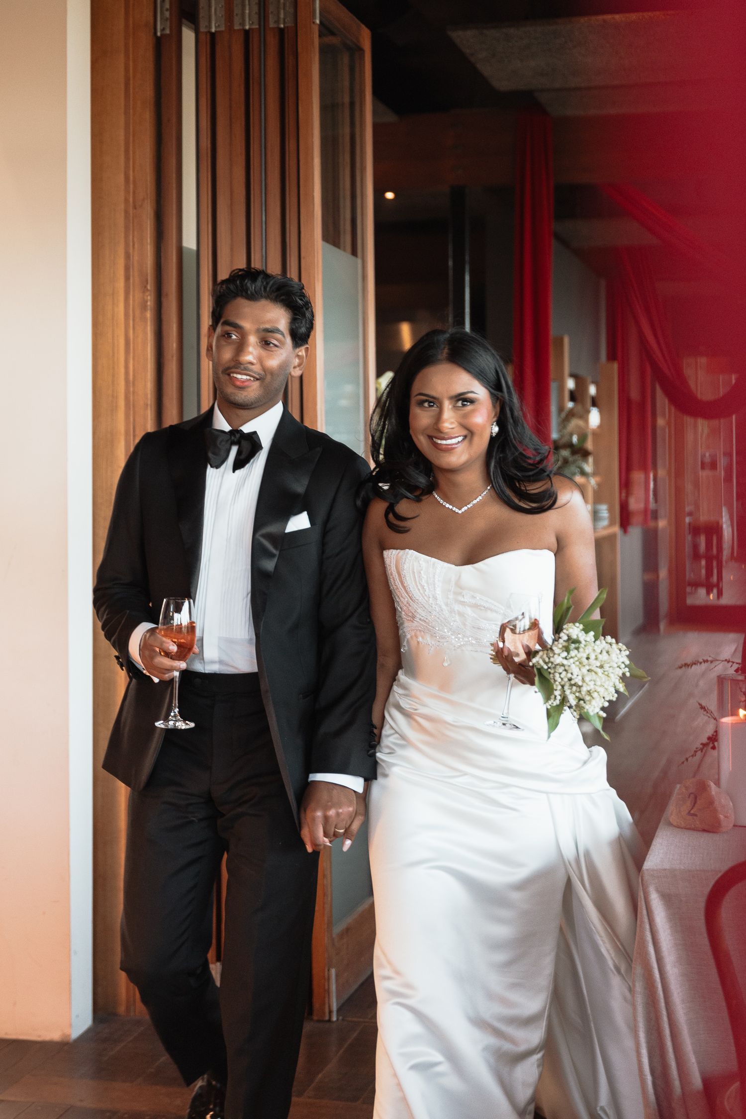 Elegant wedding couple in formal attire share champagne and laughter in a warmly lit indoor setting.