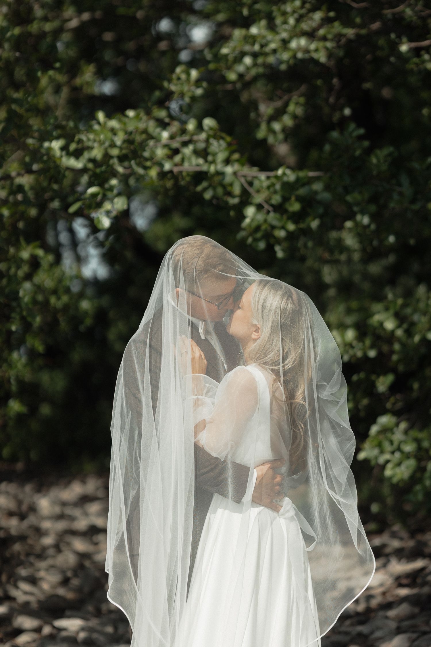 A romantic wedding couple shares an intimate moment under a flowing white veil in a garden setting.
