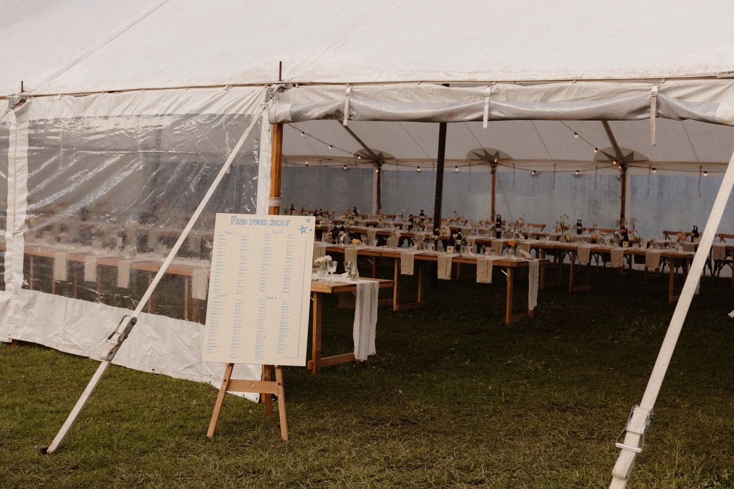 A large white event tent set up with tables and chairs inside, featuring a wooden easel at the entrance.