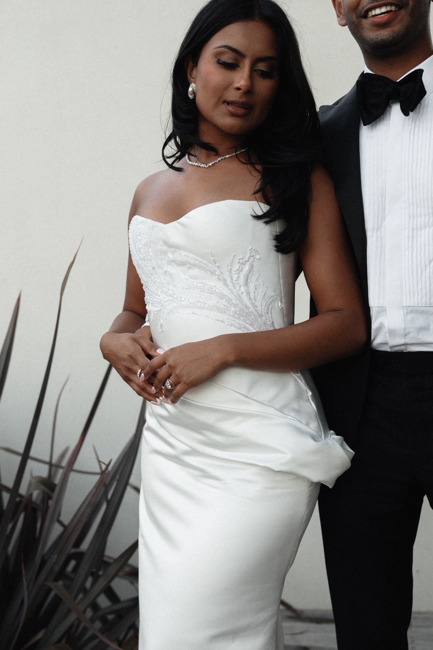 A couple in formal wedding attire poses against a white wall with dark plants in the background.