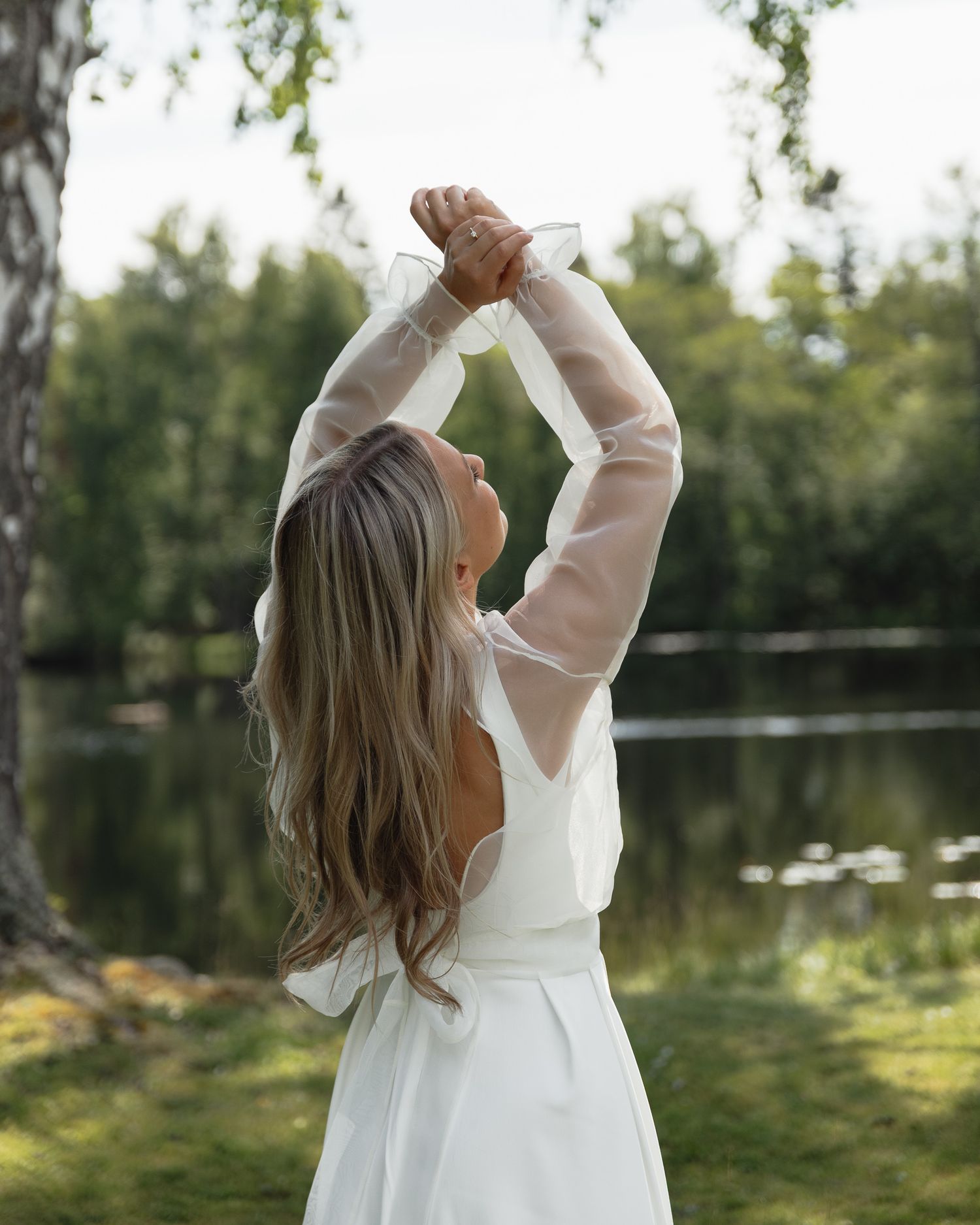 A young child in a white dress stands near a peaceful lake, reaching up towards the sky at golden hour.
