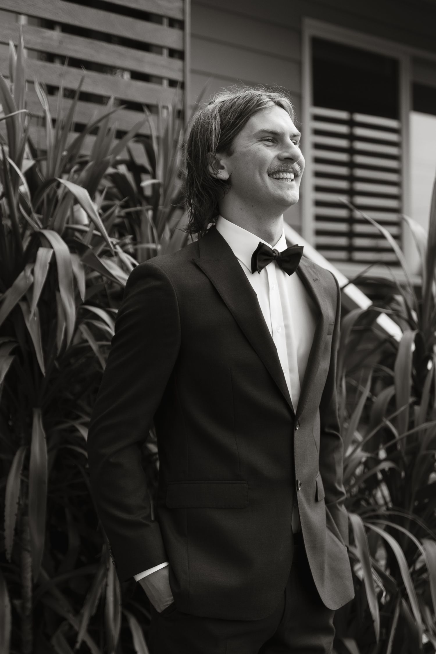 A black and white portrait of someone in a tuxedo and bowtie smiling outdoors with foliage in the background.