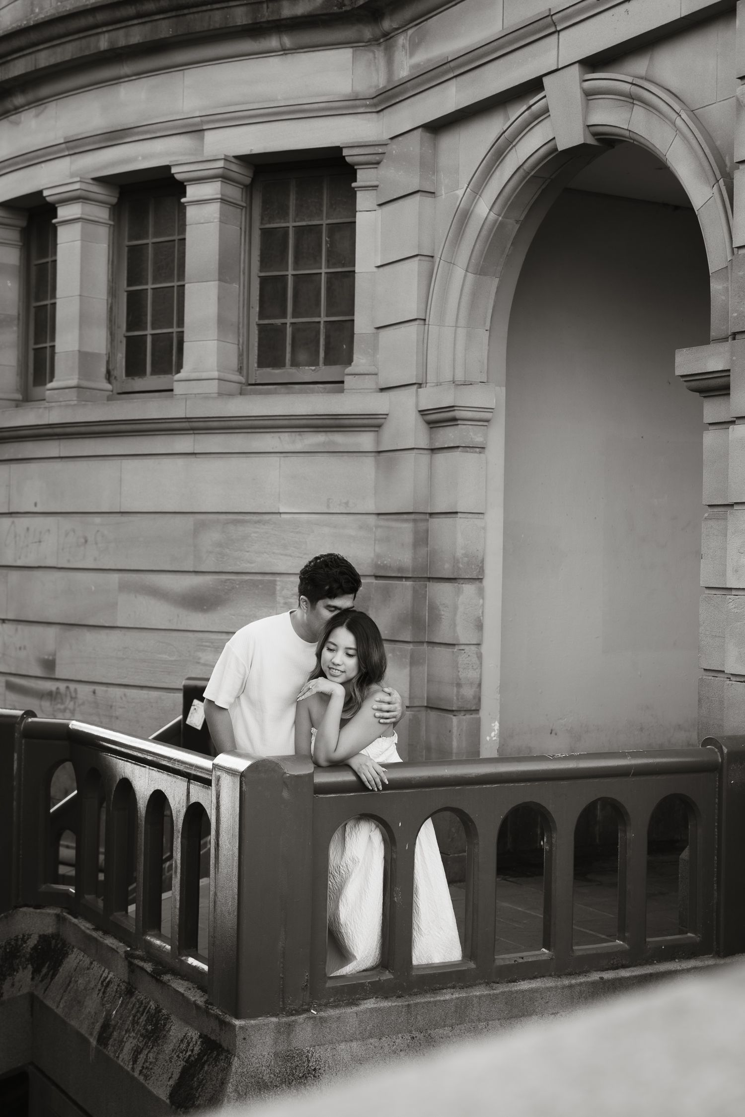 A romantic black and white photograph of a couple embracing on a balcony of a classic stone building with arched architecture.
