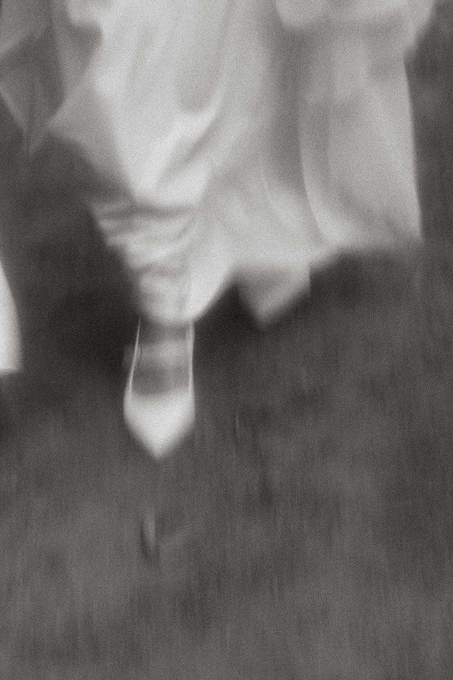 A blurred black and white image shows the bottom portion of a white dress and a high heel shoe against a dark ground.