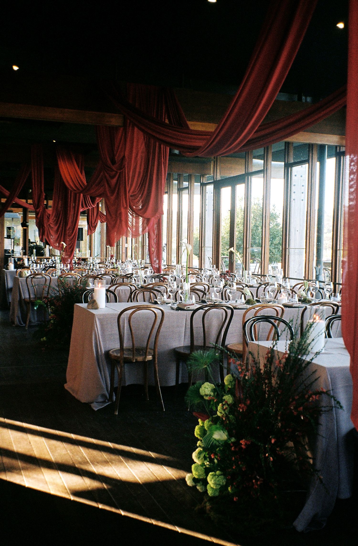 Elegant wedding reception venue with draped red curtains, round tables, and natural light streaming through large windows.