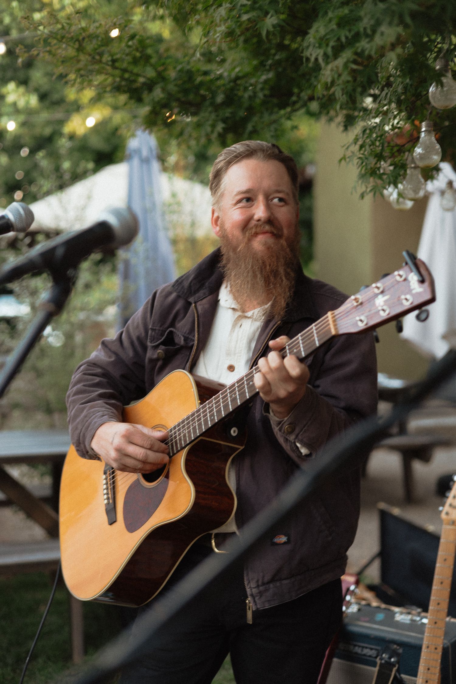 Acoustic guitarist performs outdoors on stage with a wooden guitar against a natural green backdrop.