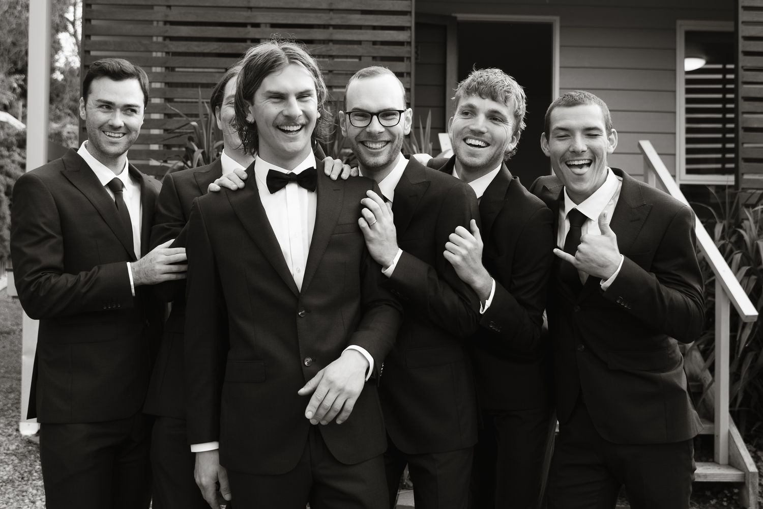 A black and white photo shows five groomsmen in formal suits and bowties posing together on outdoor steps.