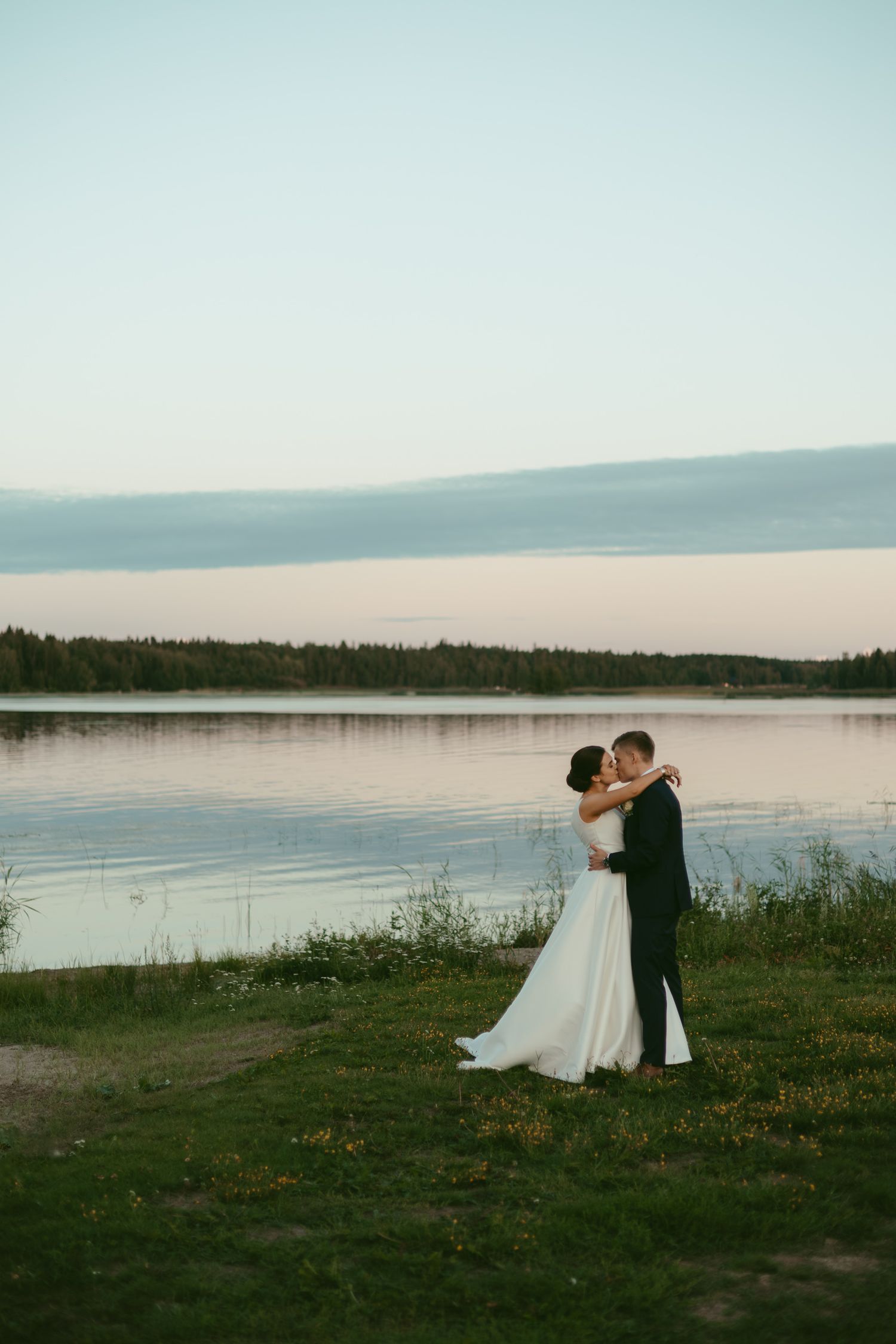 Newlyweds embrace by a serene lake at sunset with mountains in the distance.