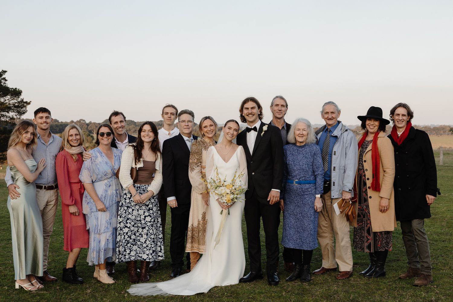 Large group photo of wedding guests and newlyweds on a grassy outdoor venue at dusk.
