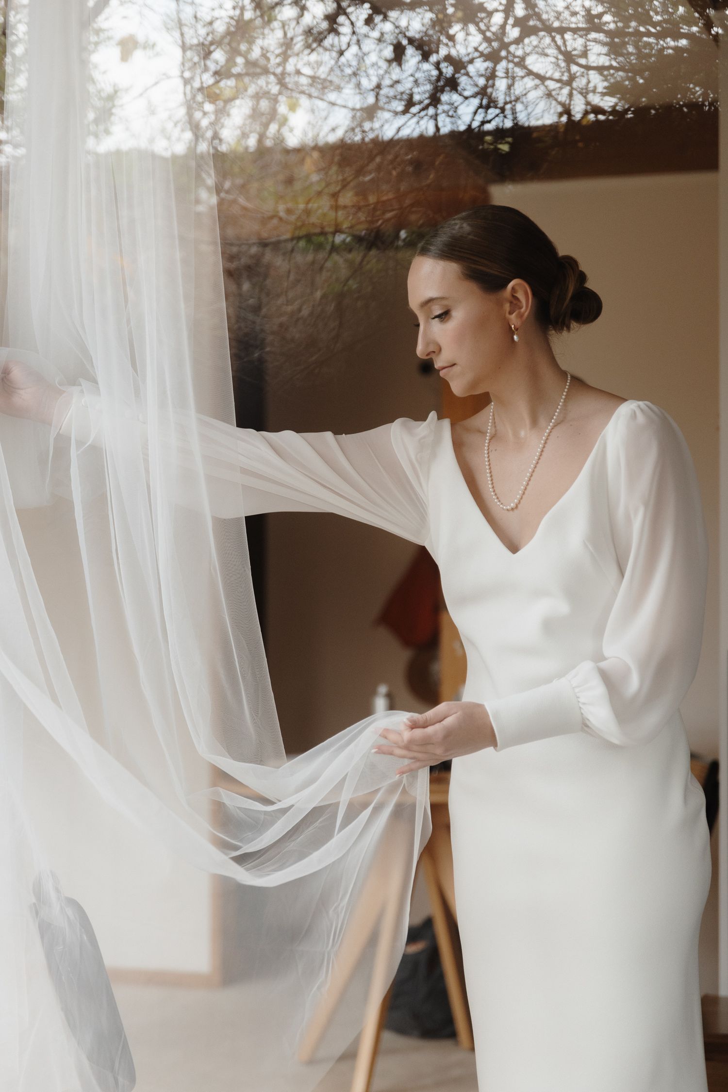 Woman in sleek white wedding dress with flowing sleeves adjusts veil near window.