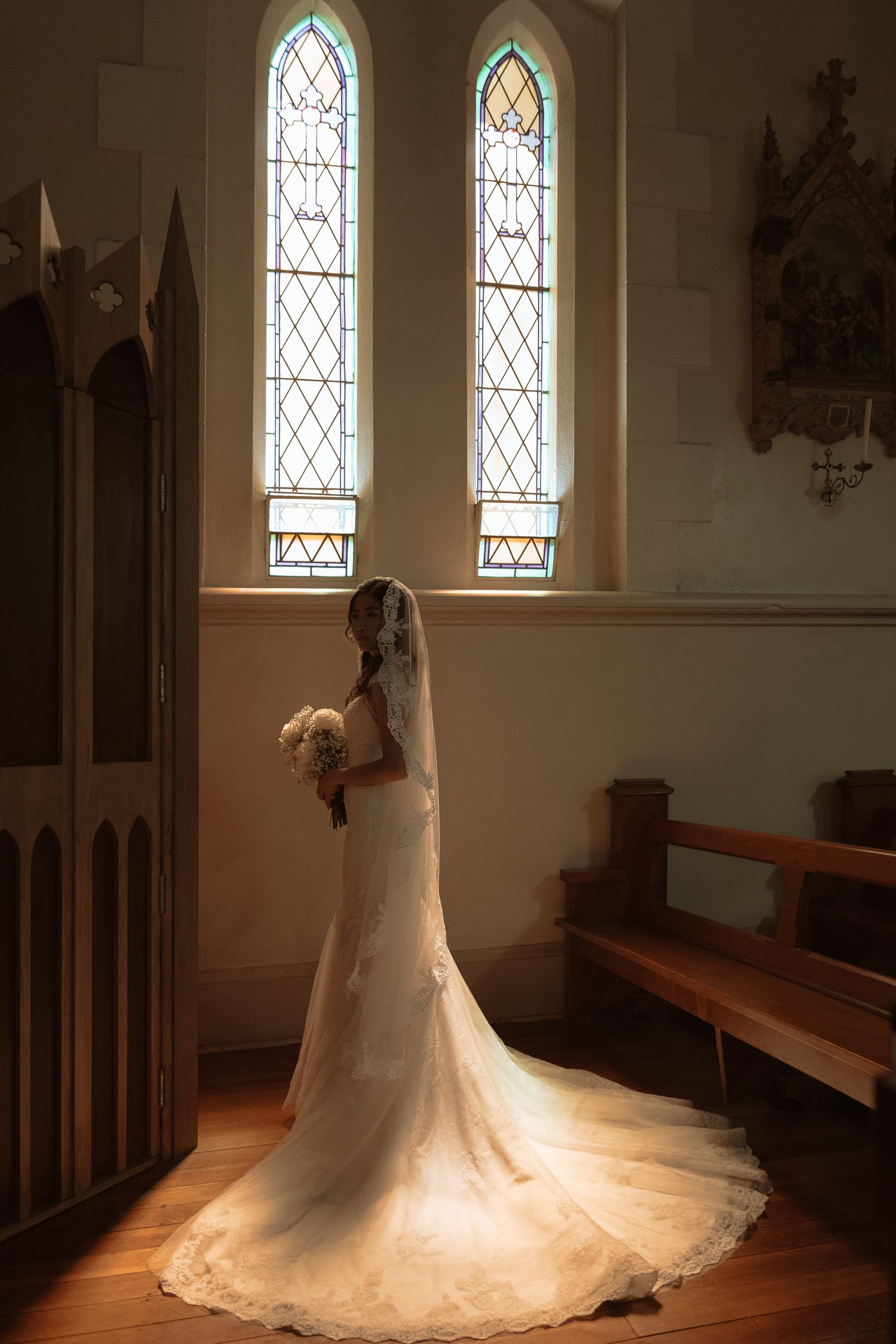 Bride in lace gown stands in dramatic sunlight streaming through stained glass windows.