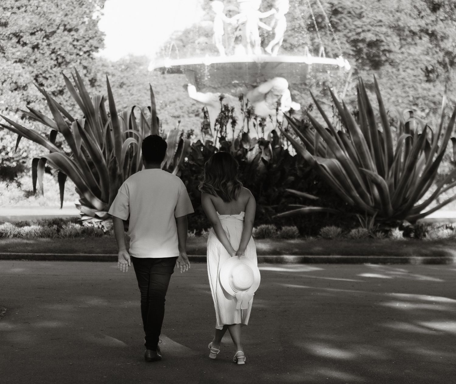 A couple walks hand in hand down a shaded garden path with large agave plants lining the walkway in black and white.