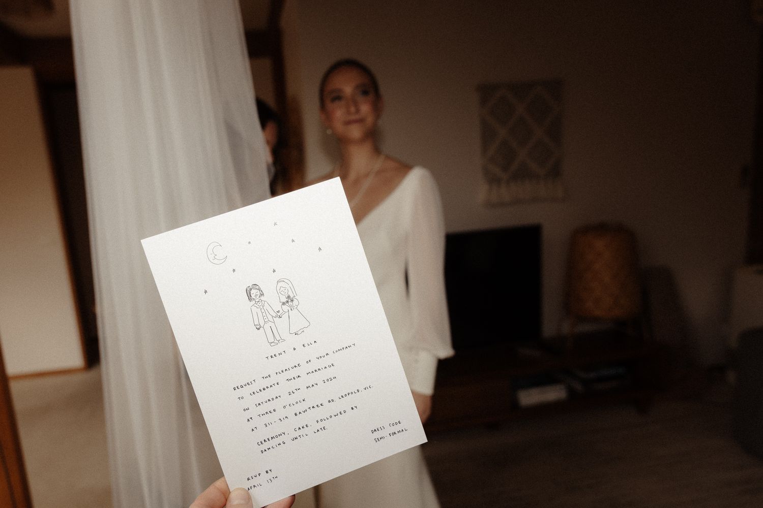 Someone in a white v-neck dress holds up a wedding invitation with handwritten text in a dimly lit room.