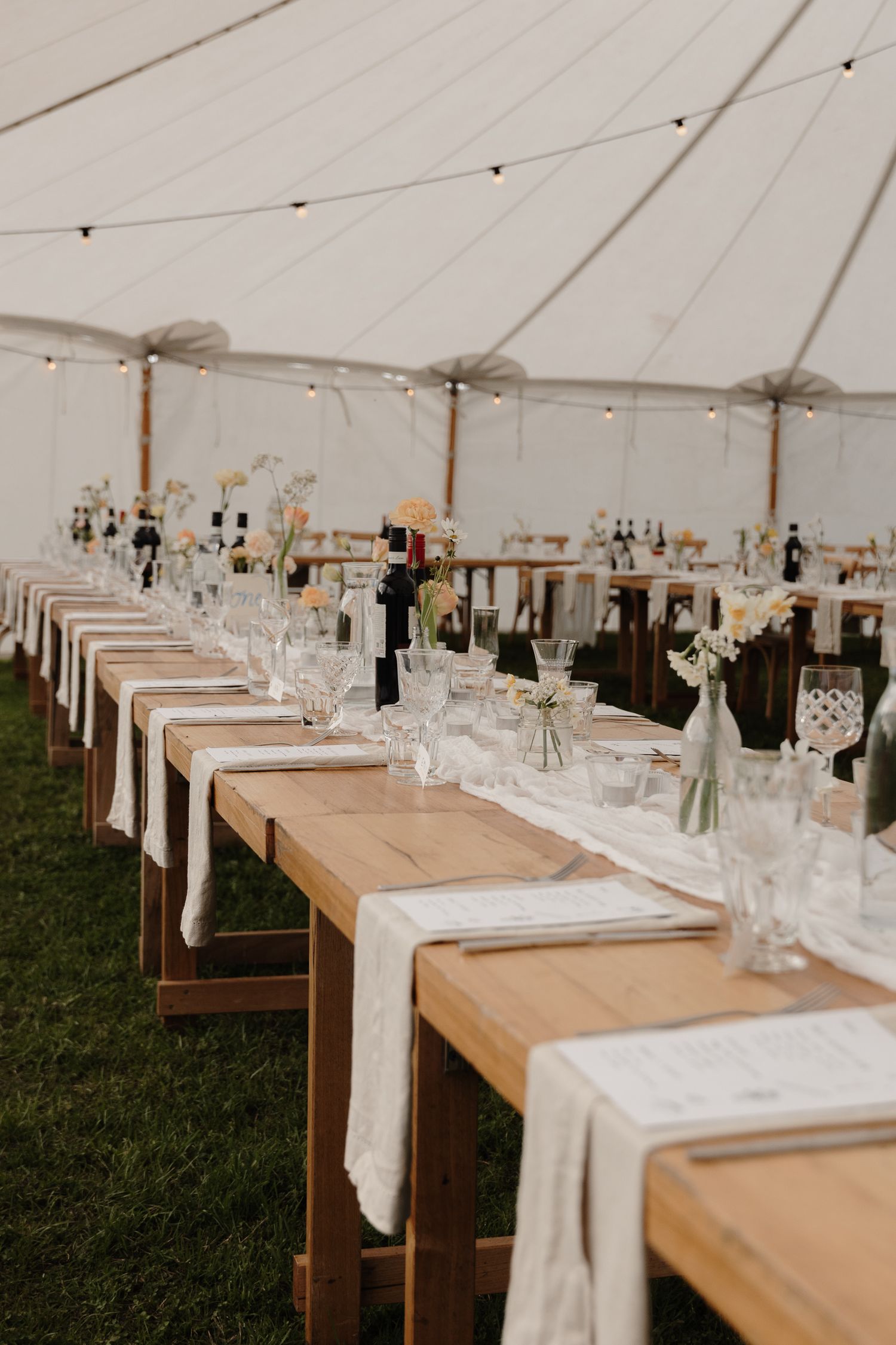 Rustic wooden farm tables with white linens and glassware set up for an elegant wedding reception under a white tent.