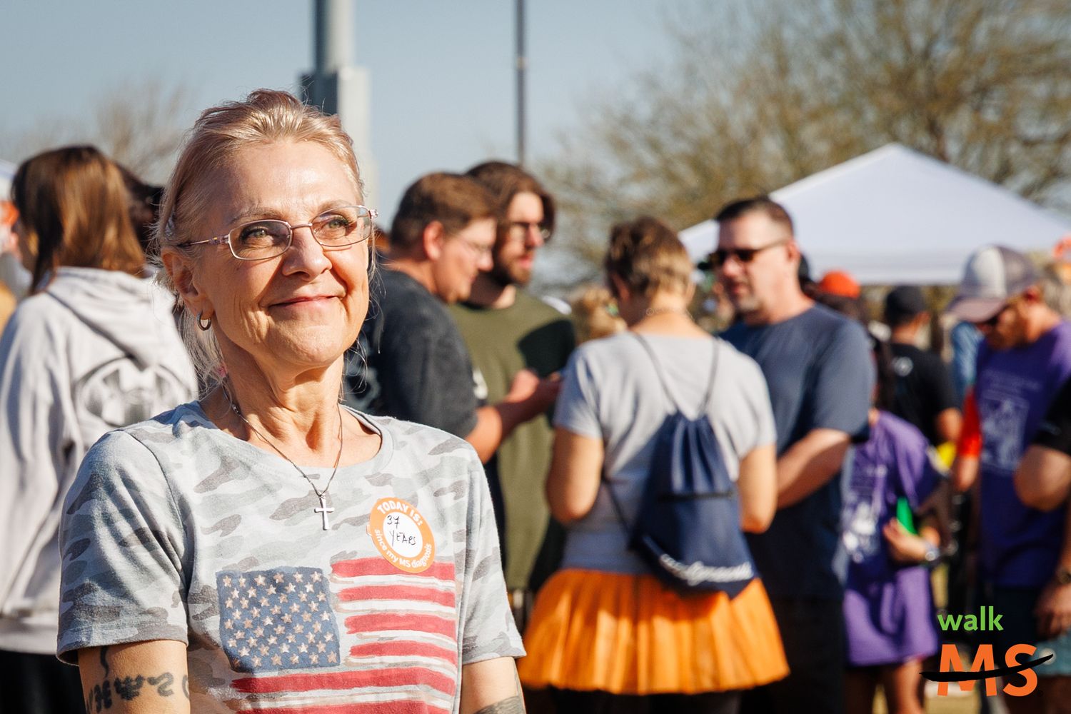Smiling woman at an outdoor event with people gathered in the background, captured during a candid moment. Event photography by Sasy Cassie.